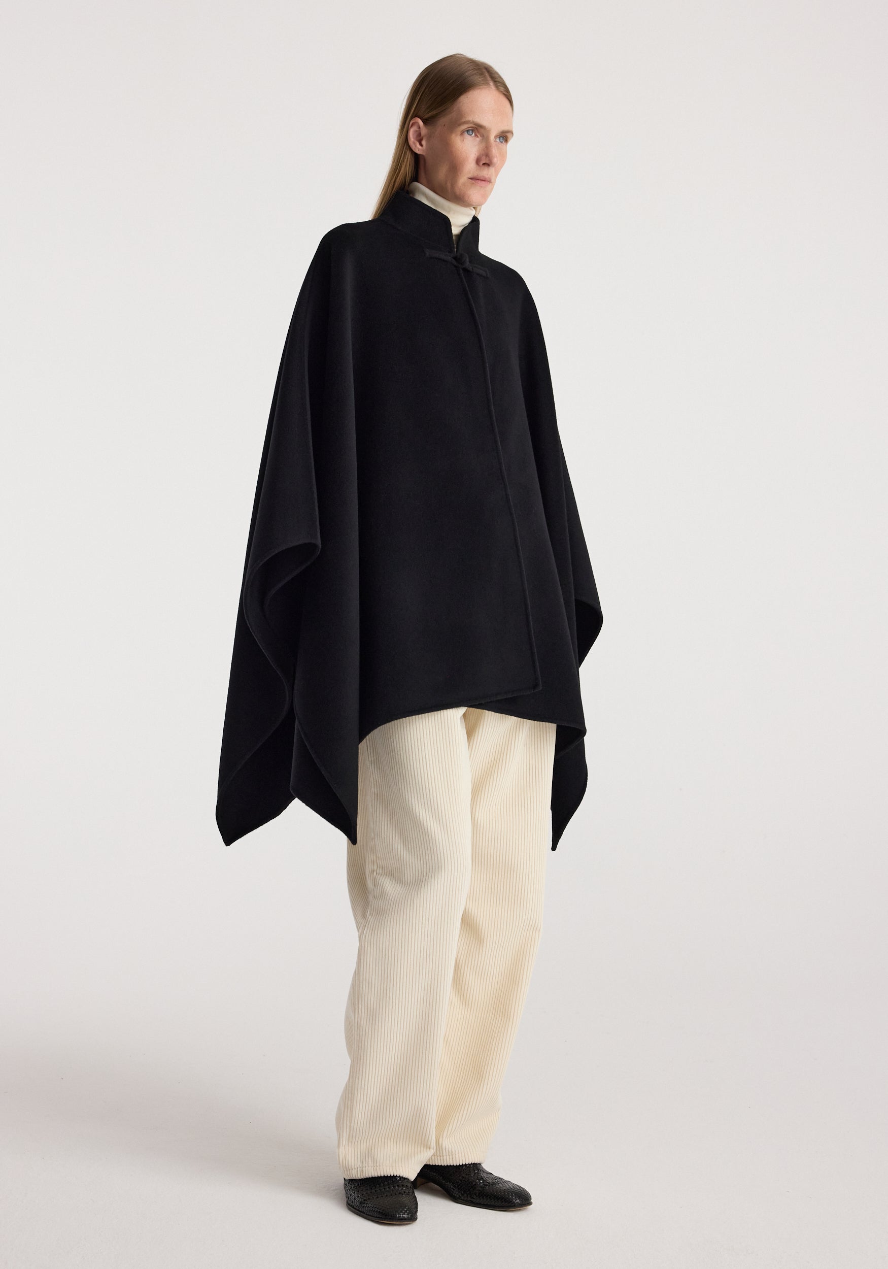Woman wearing a black Double-faced pankou poncho | black over a light turtleneck and cream pants, standing against a plain background in a relaxed pose.