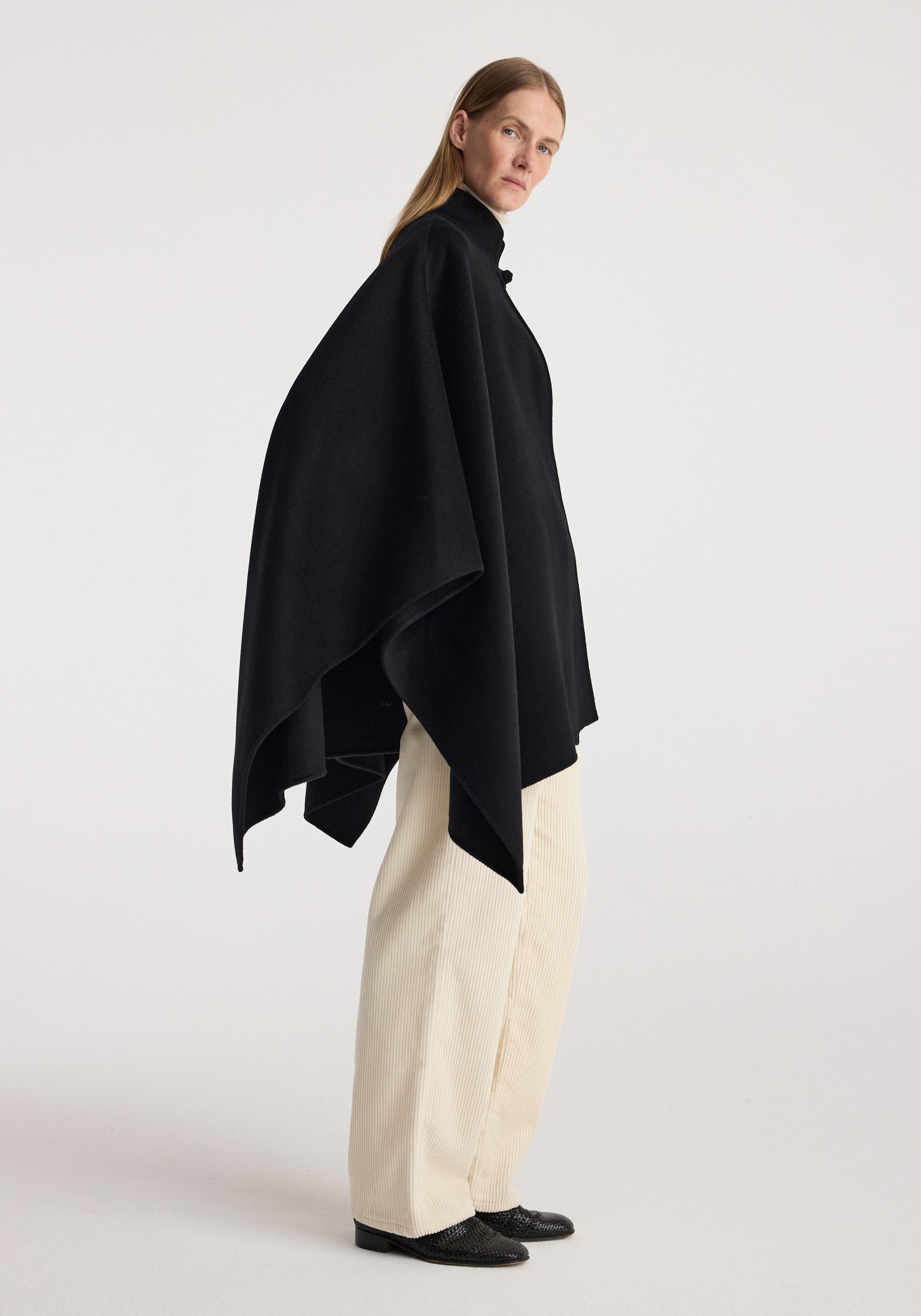 Woman wearing a black Double-faced pankou poncho | black with a high neck, paired with cream wide-leg pants and black shoes, standing against a plain background.