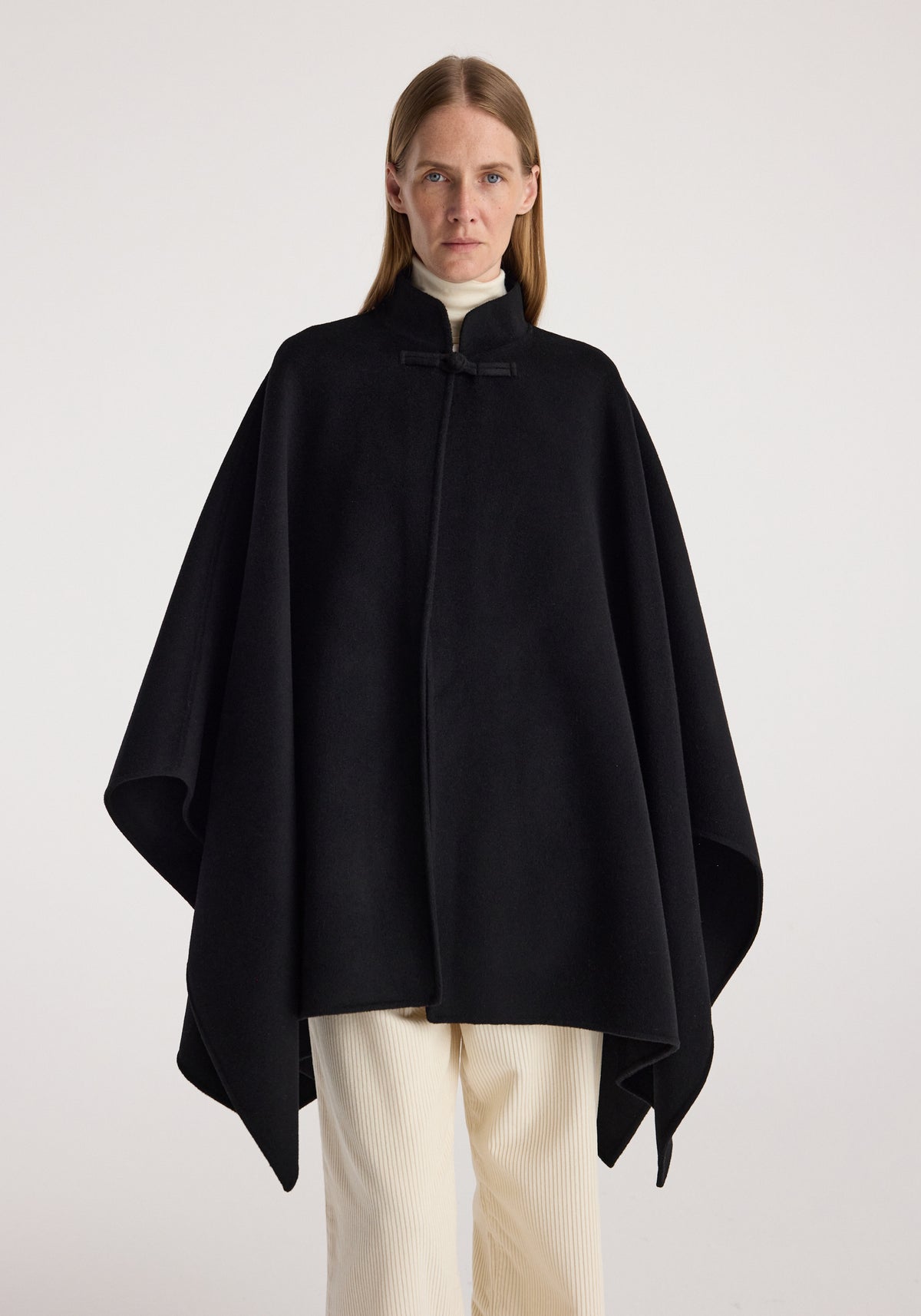 Woman wearing a black double-faced pankou poncho | black over a light turtleneck and cream pants, standing against a plain background with a serious expression.