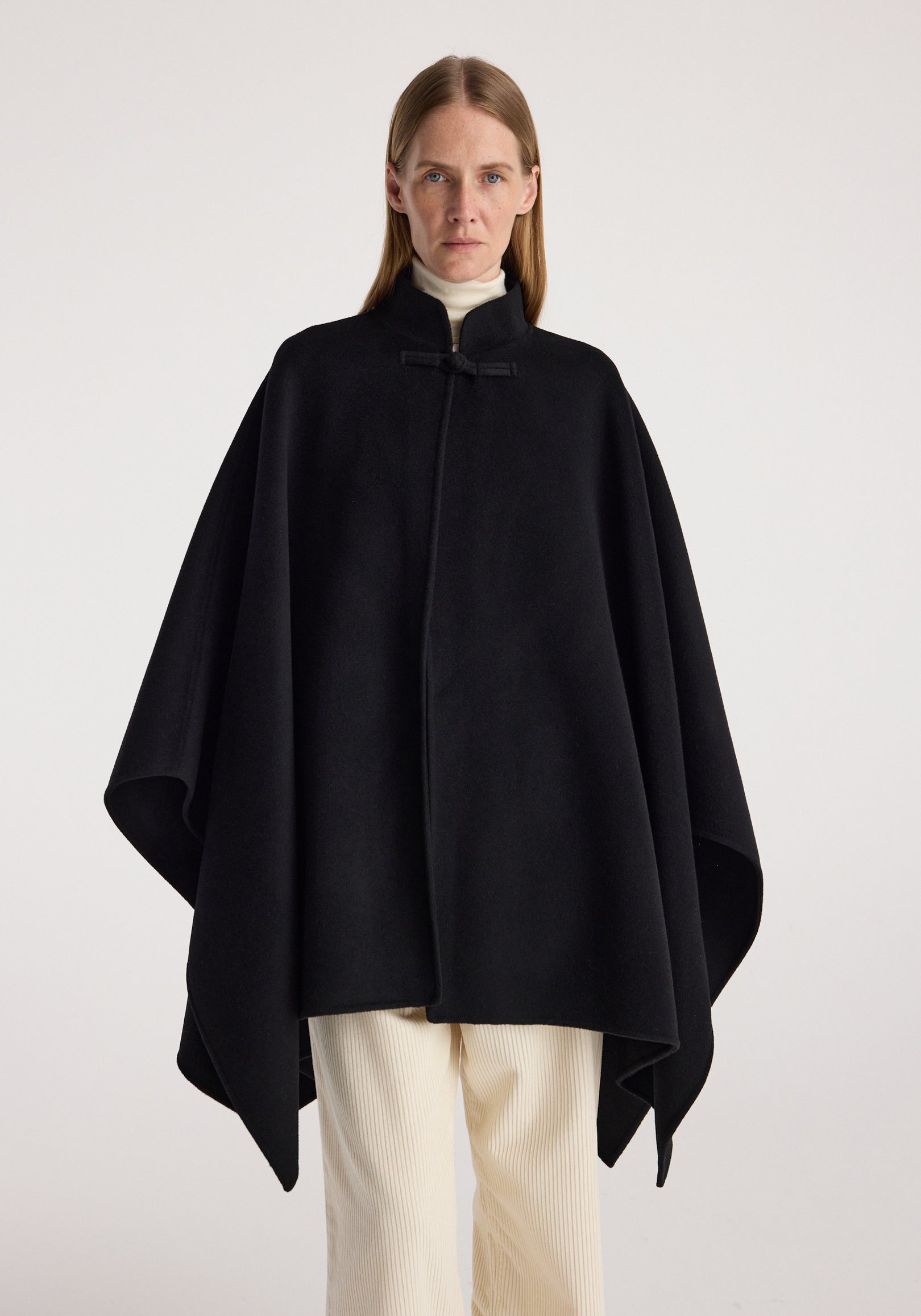 Woman wearing a black double-faced pankou poncho | black over a light turtleneck and cream pants, standing against a plain background with a serious expression.