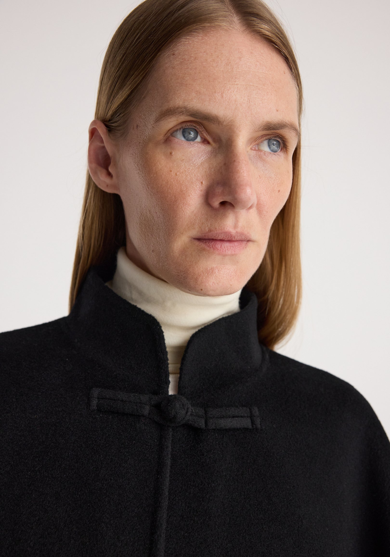 Close-up of a woman wearing a black Double-faced pankou poncho | black with a high collar and traditional knot button detail over a white turtleneck.