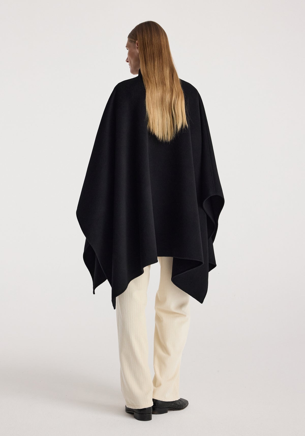 Woman seen from behind wearing a black Double-faced pankou poncho | black over white pants and black shoes against a plain light background