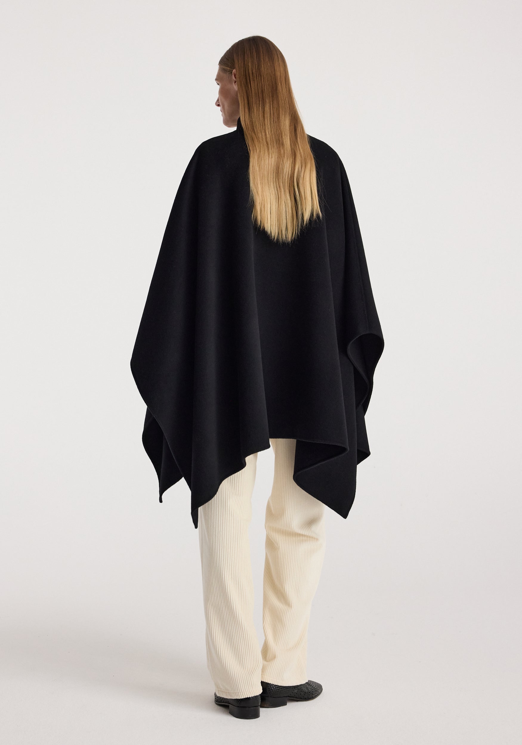 Woman seen from behind wearing a black Double-faced pankou poncho | black over white pants and black shoes against a plain light background