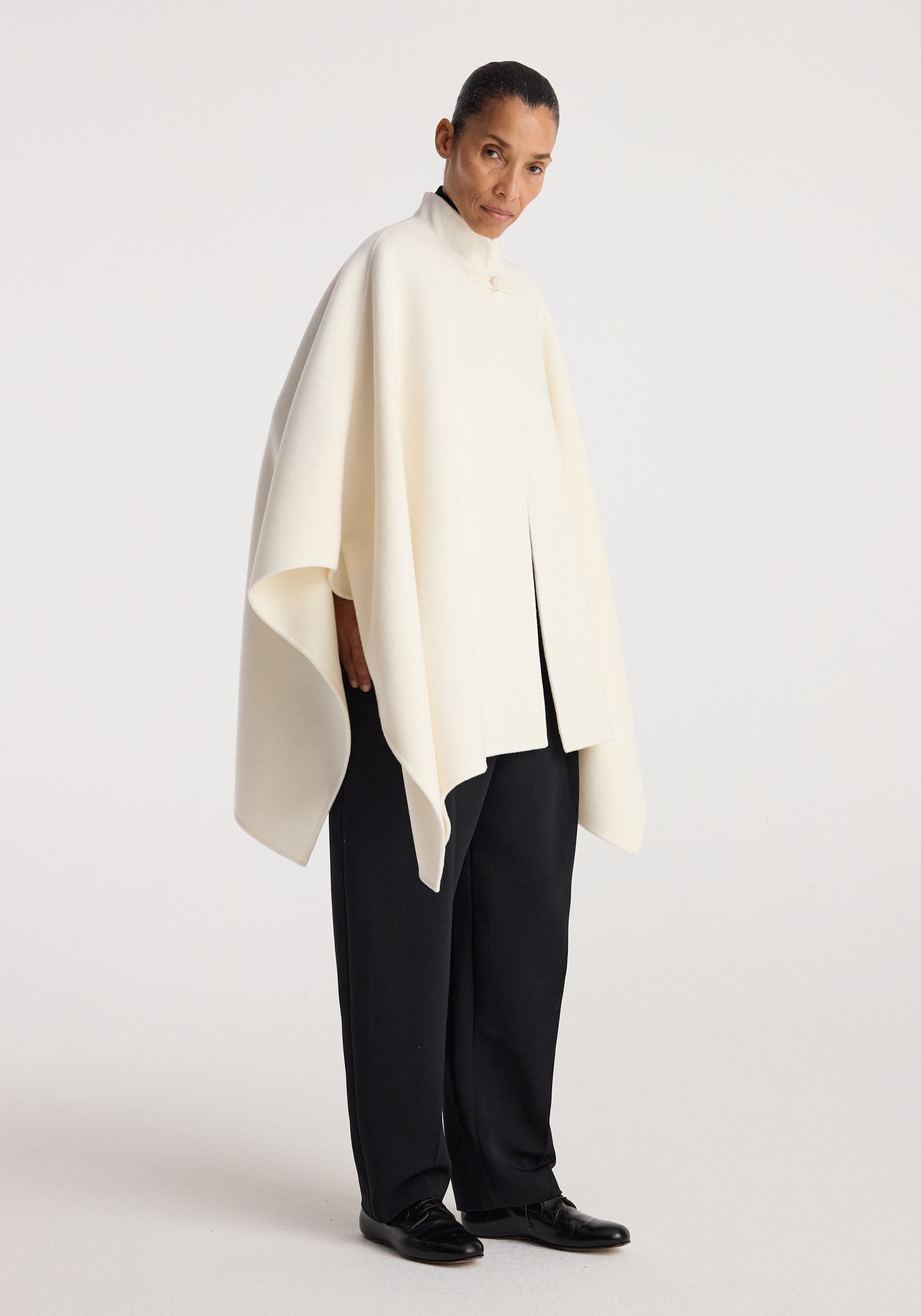 Woman wearing an off white double-faced pankou poncho with a high collar and loose fit, paired with black wide-leg trousers and black shoes on a plain background.