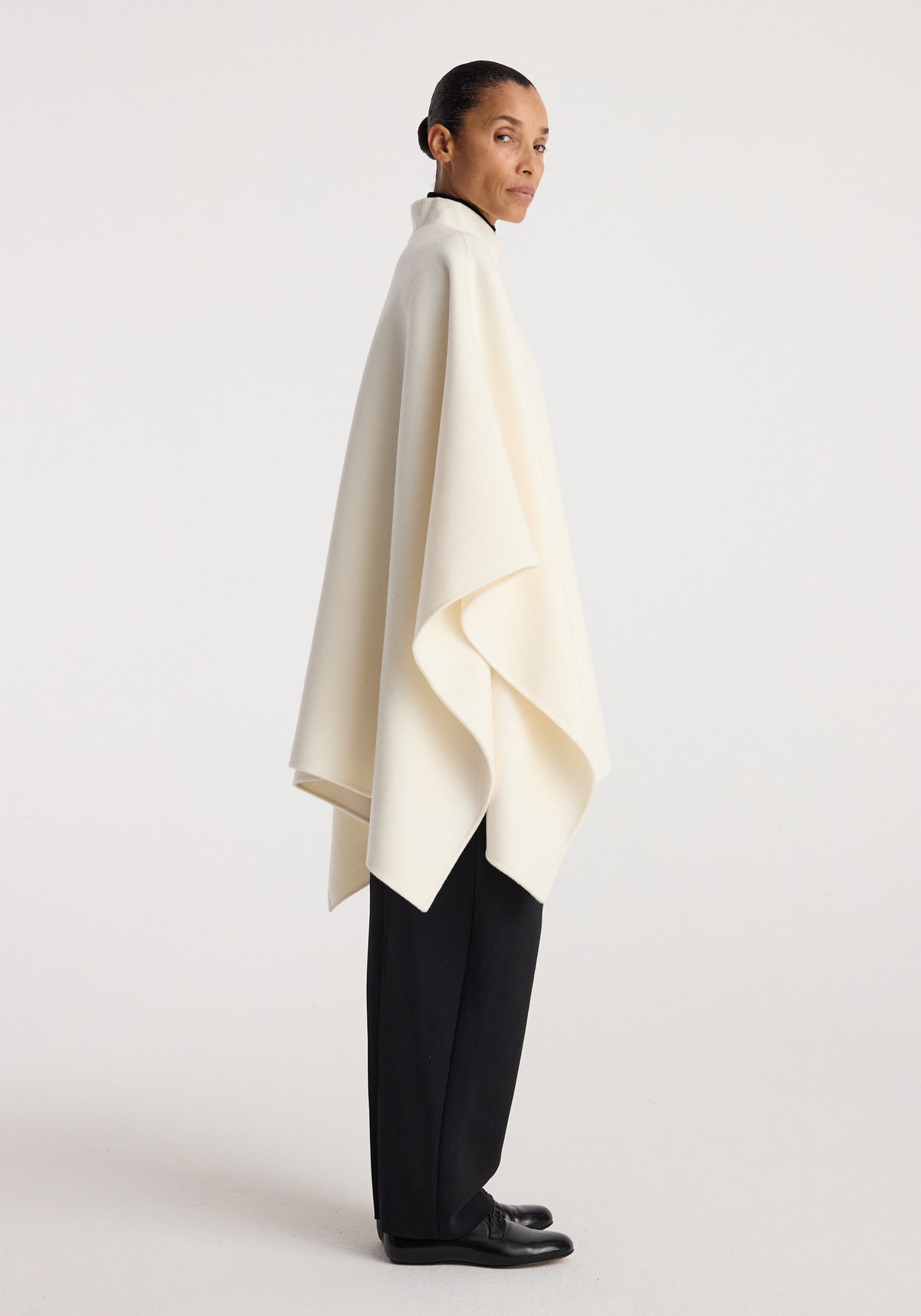 Woman wearing a sleek off white double-faced pankou poncho over black trousers and shoes, standing in a side profile against a plain background, showcasing the poncho's elegant drape and clean lines.