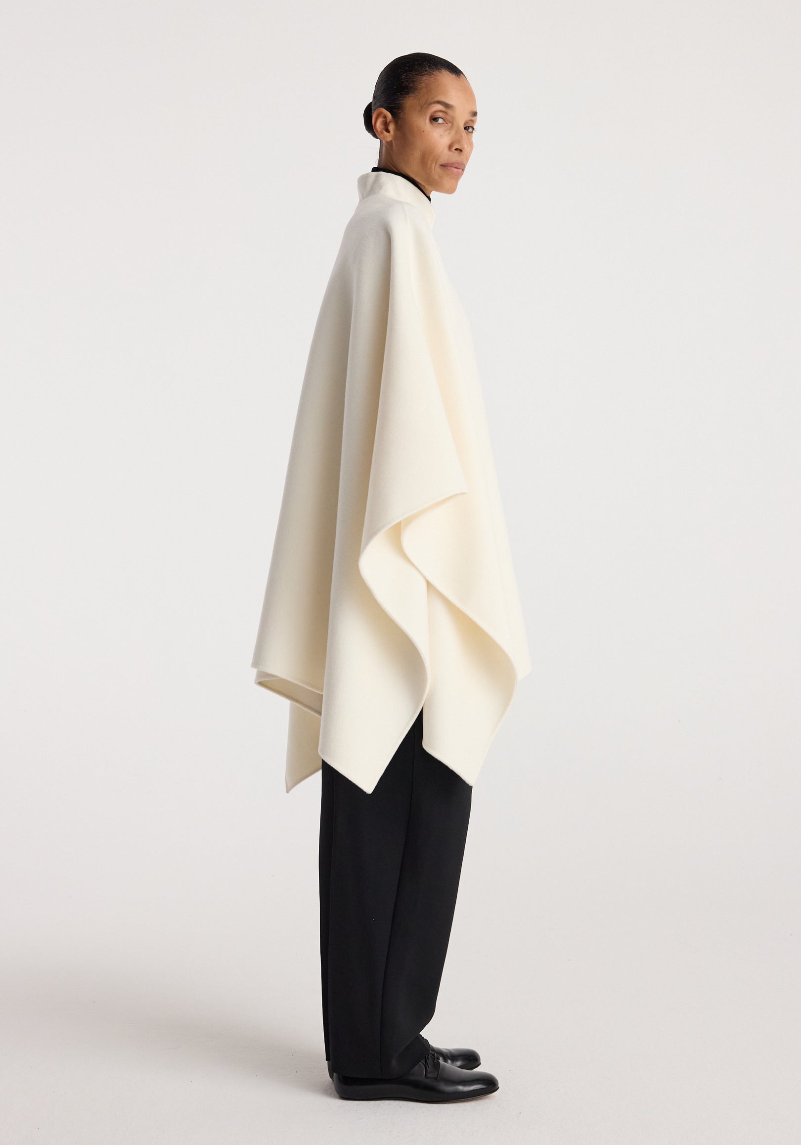 Woman wearing a sleek off white double-faced pankou poncho over black trousers and shoes, standing in a side profile against a plain background, showcasing the poncho's elegant drape and clean lines.