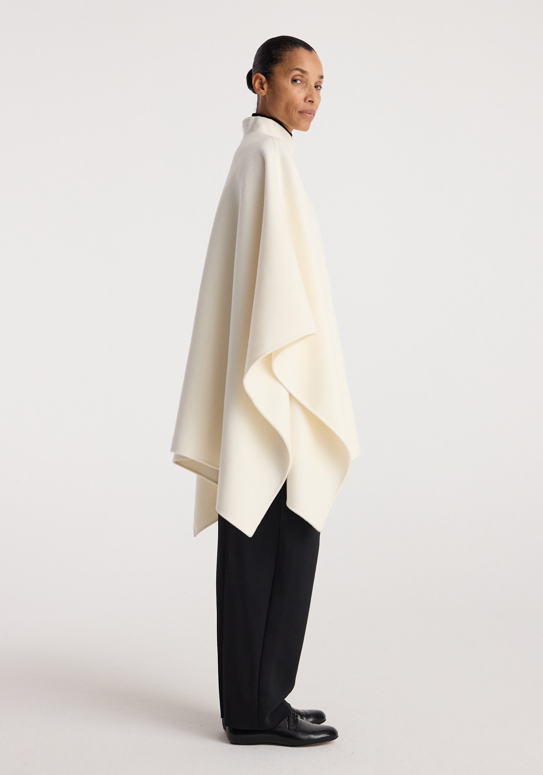 Woman wearing a sleek off white double-faced pankou poncho over black trousers and shoes, standing in a side profile against a plain background, showcasing the poncho's elegant drape and clean lines.