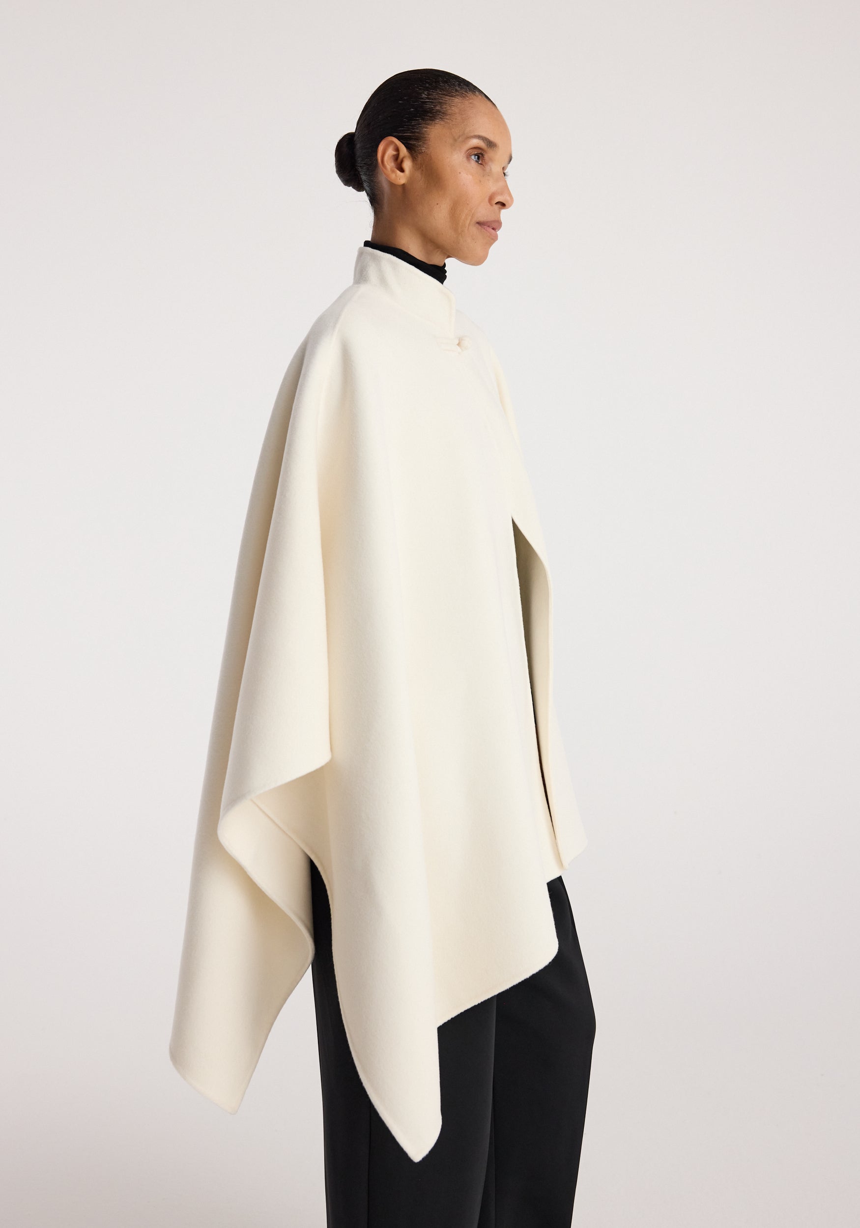 Profile view of a woman wearing a minimalist off-white Double-faced pankou poncho | off white over a black outfit against a plain light background.