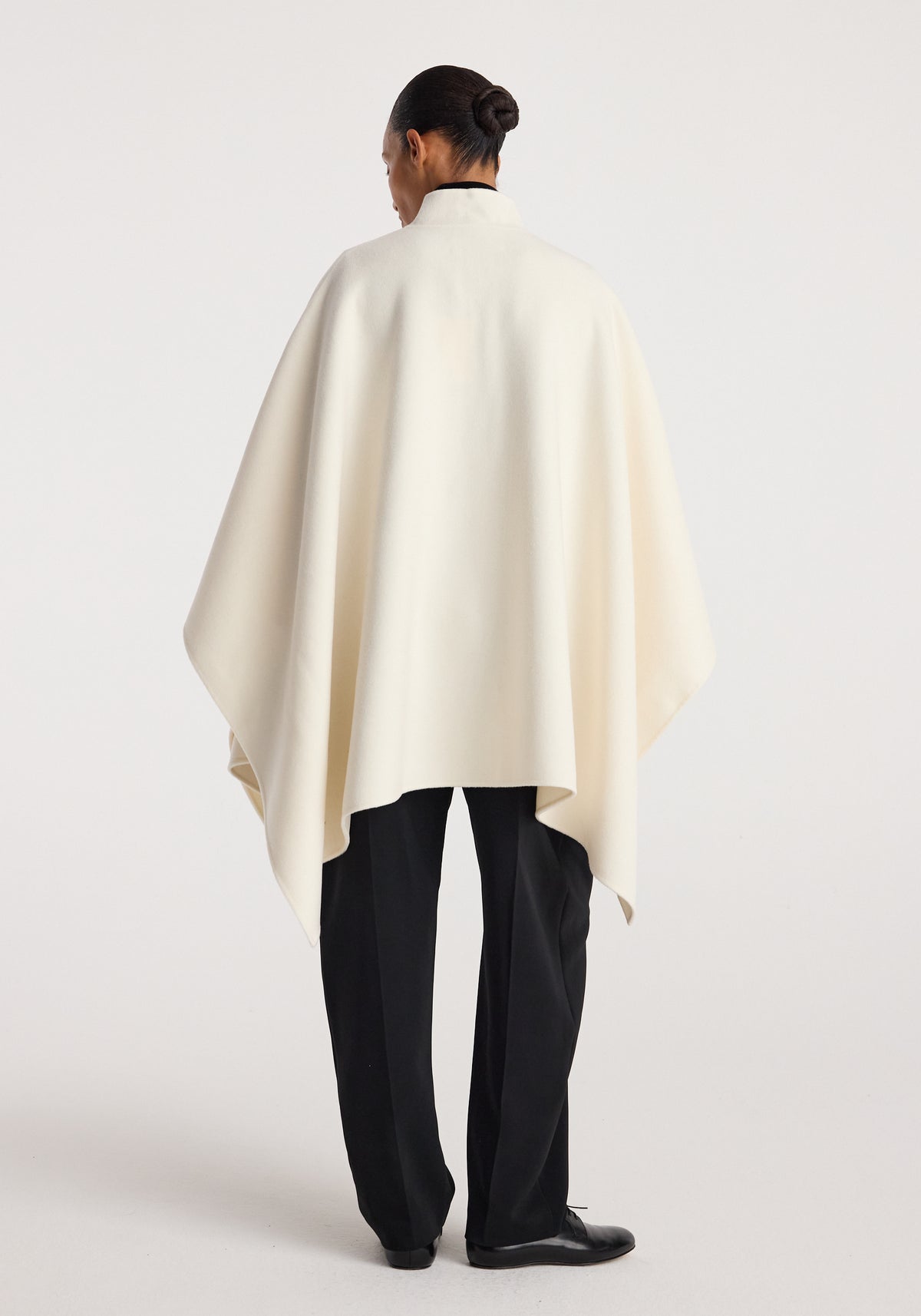 Rear view of a person wearing a flowing off white Double-faced pankou poncho | off white over black trousers, standing against a plain background.