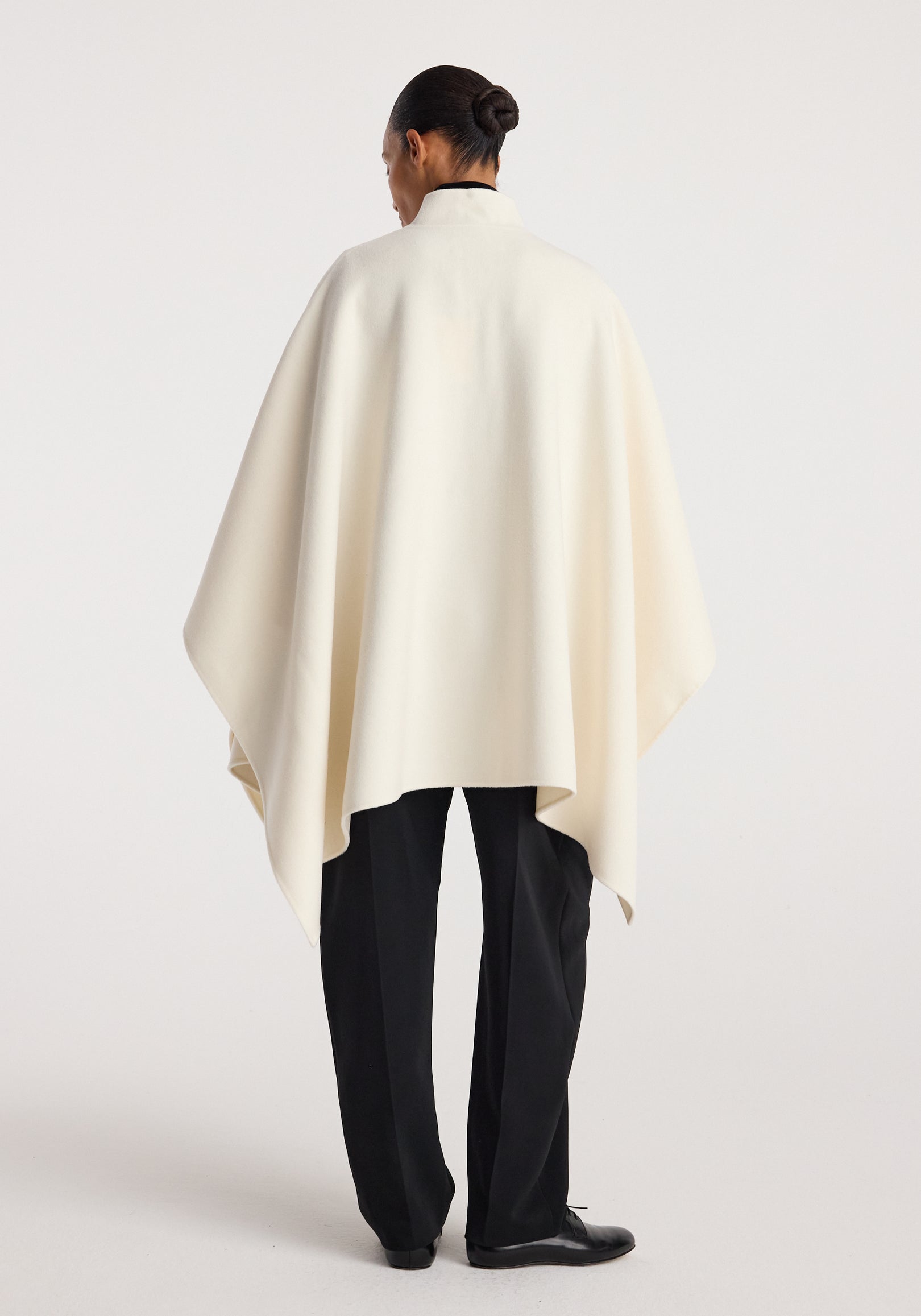 Rear view of a person wearing a flowing off white Double-faced pankou poncho | off white over black trousers, standing against a plain background.