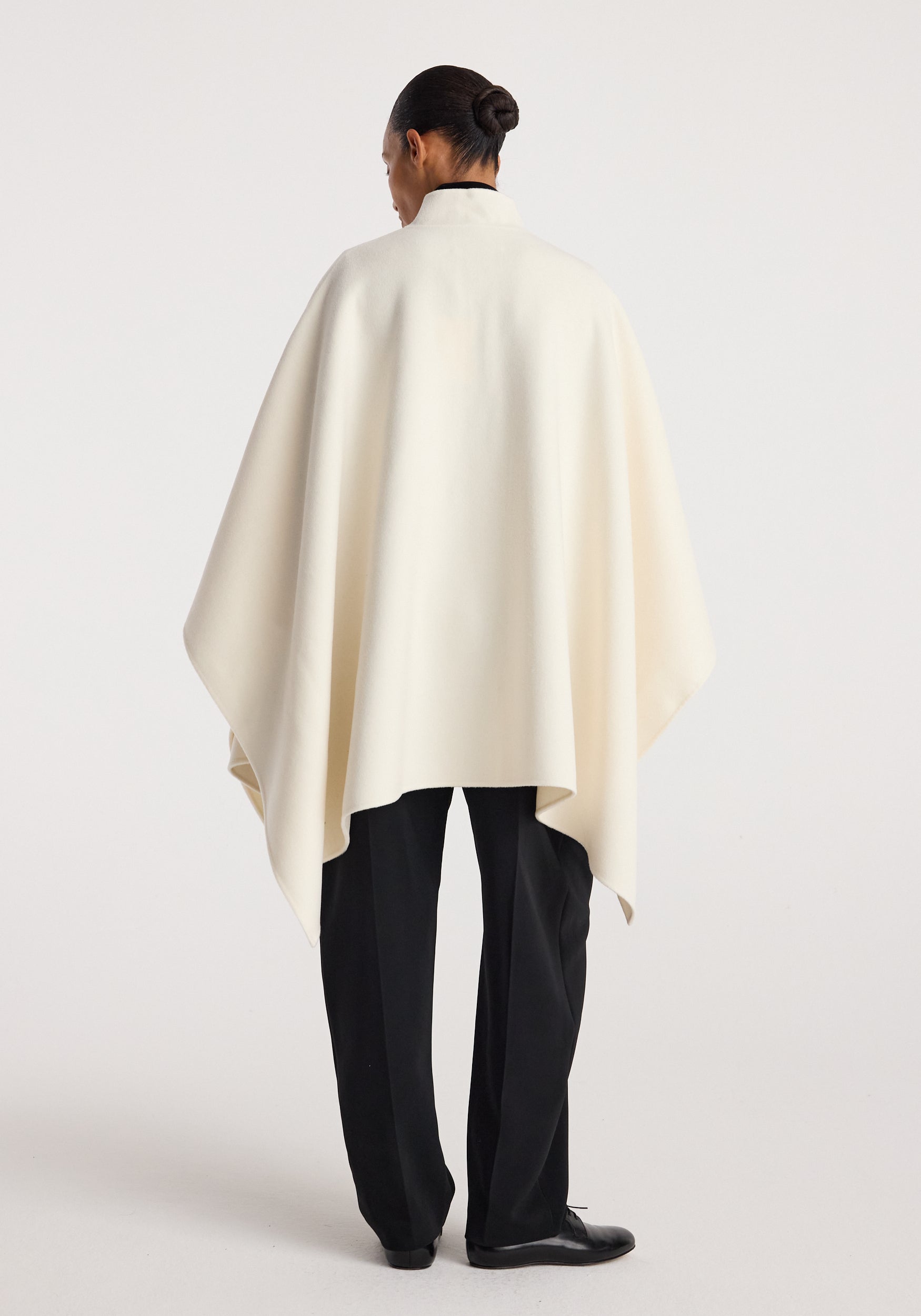Rear view of a person wearing a flowing off white Double-faced pankou poncho | off white over black trousers, standing against a plain background.
