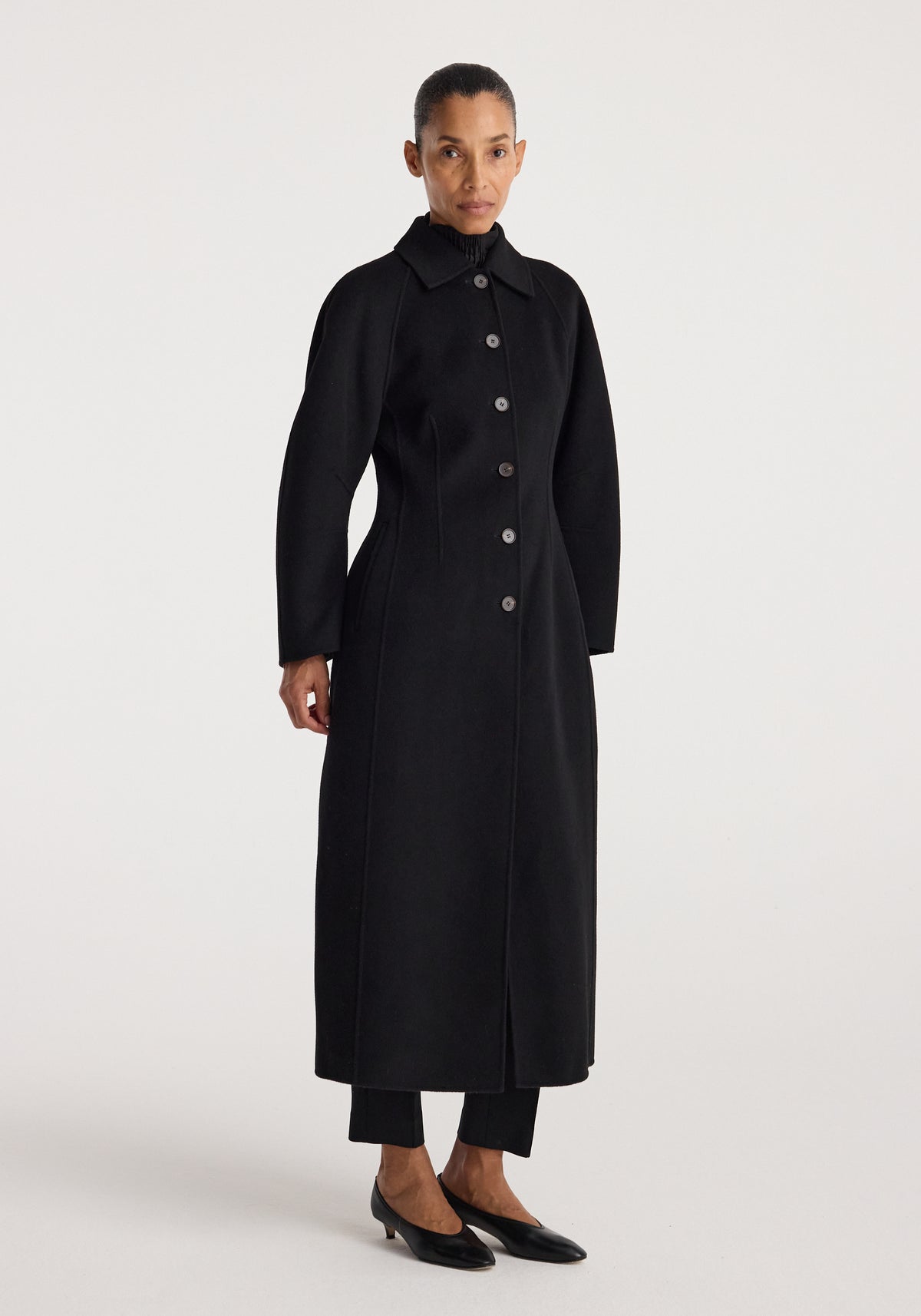 Woman wearing a minimalist black Double-faced elongated sculptural coat | black with a button-up front and a high collar, standing against a plain background.