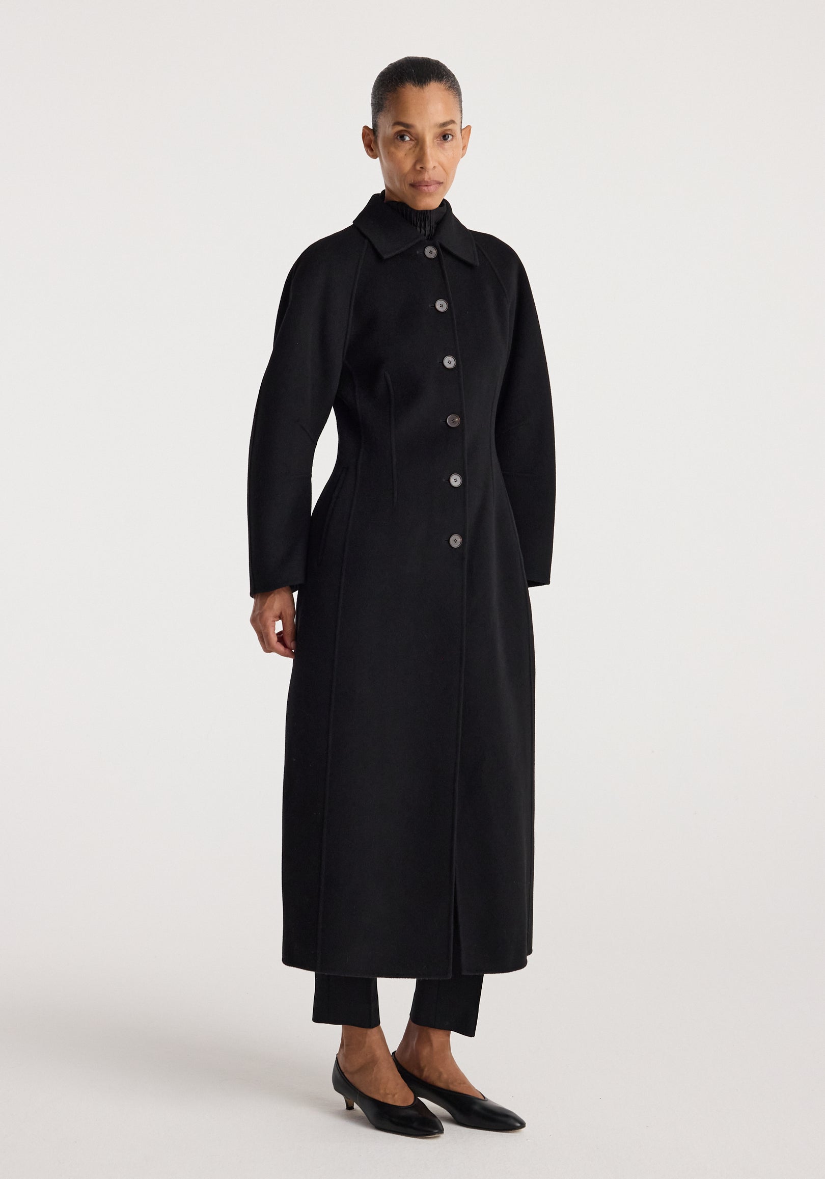 Woman wearing a minimalist black Double-faced elongated sculptural coat | black with a button-up front and a high collar, standing against a plain background.