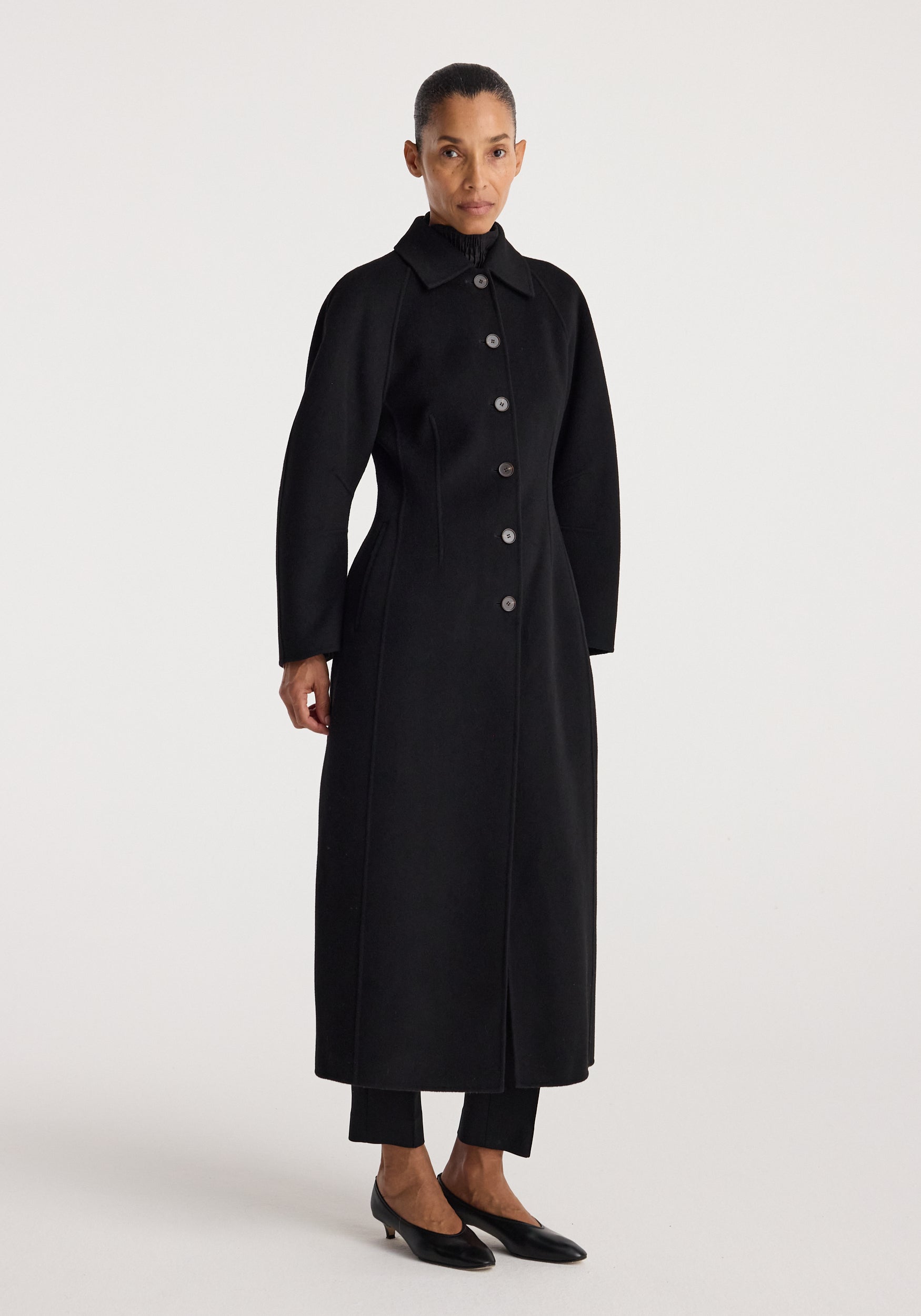 Woman wearing a minimalist black Double-faced elongated sculptural coat | black with a button-up front and a high collar, standing against a plain background.