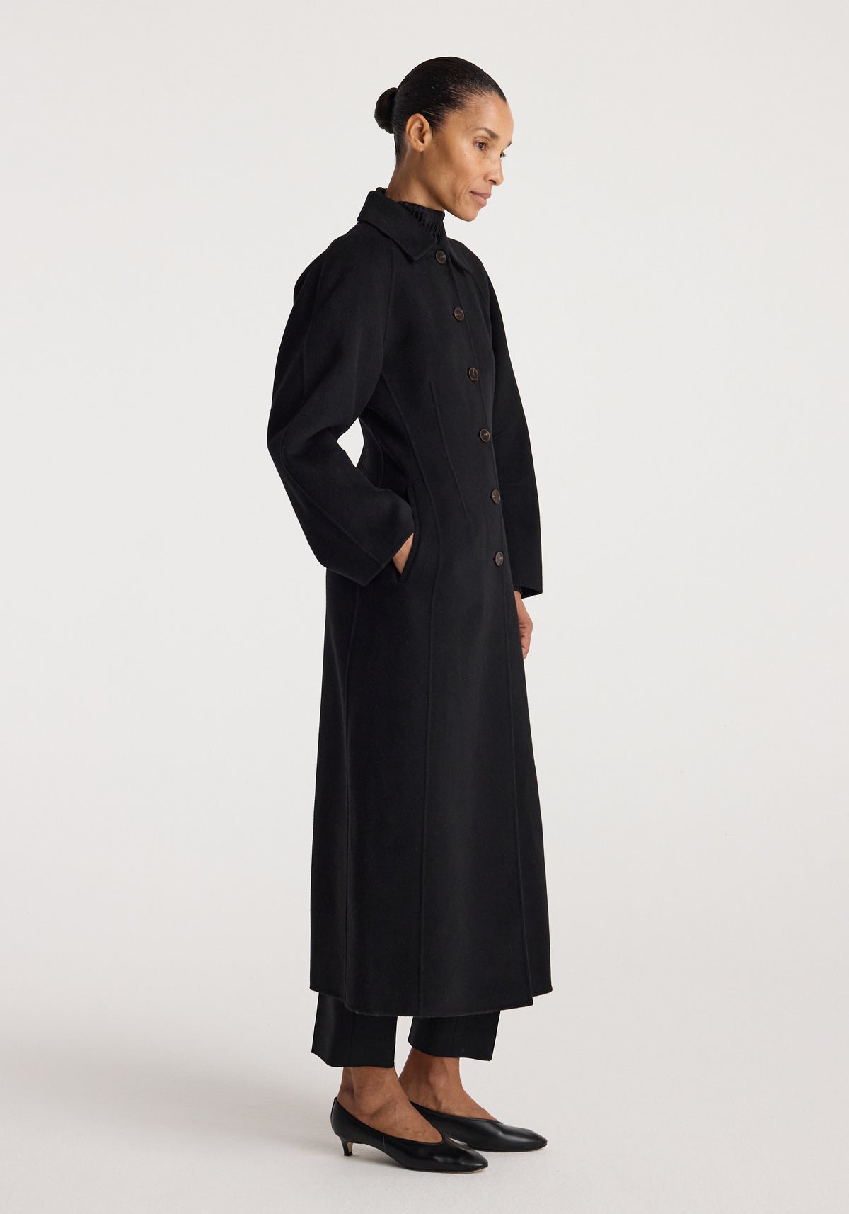 Woman wearing a long black Double-faced elongated sculptural coat | black with button closure, side pockets, and a structured collar, styled with black pants and low heels.