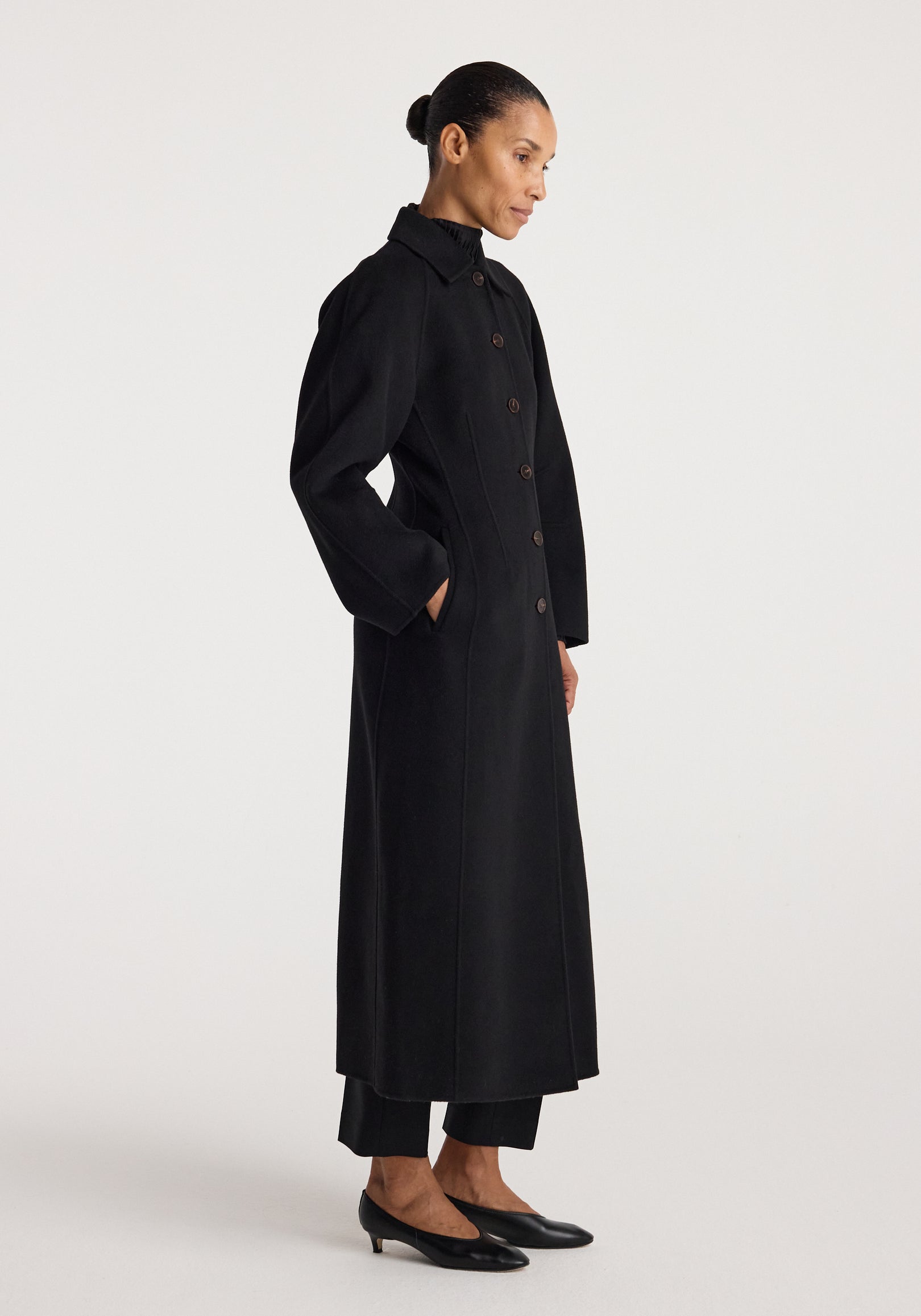 Woman wearing a long black Double-faced elongated sculptural coat | black with button closure, side pockets, and a structured collar, styled with black pants and low heels.