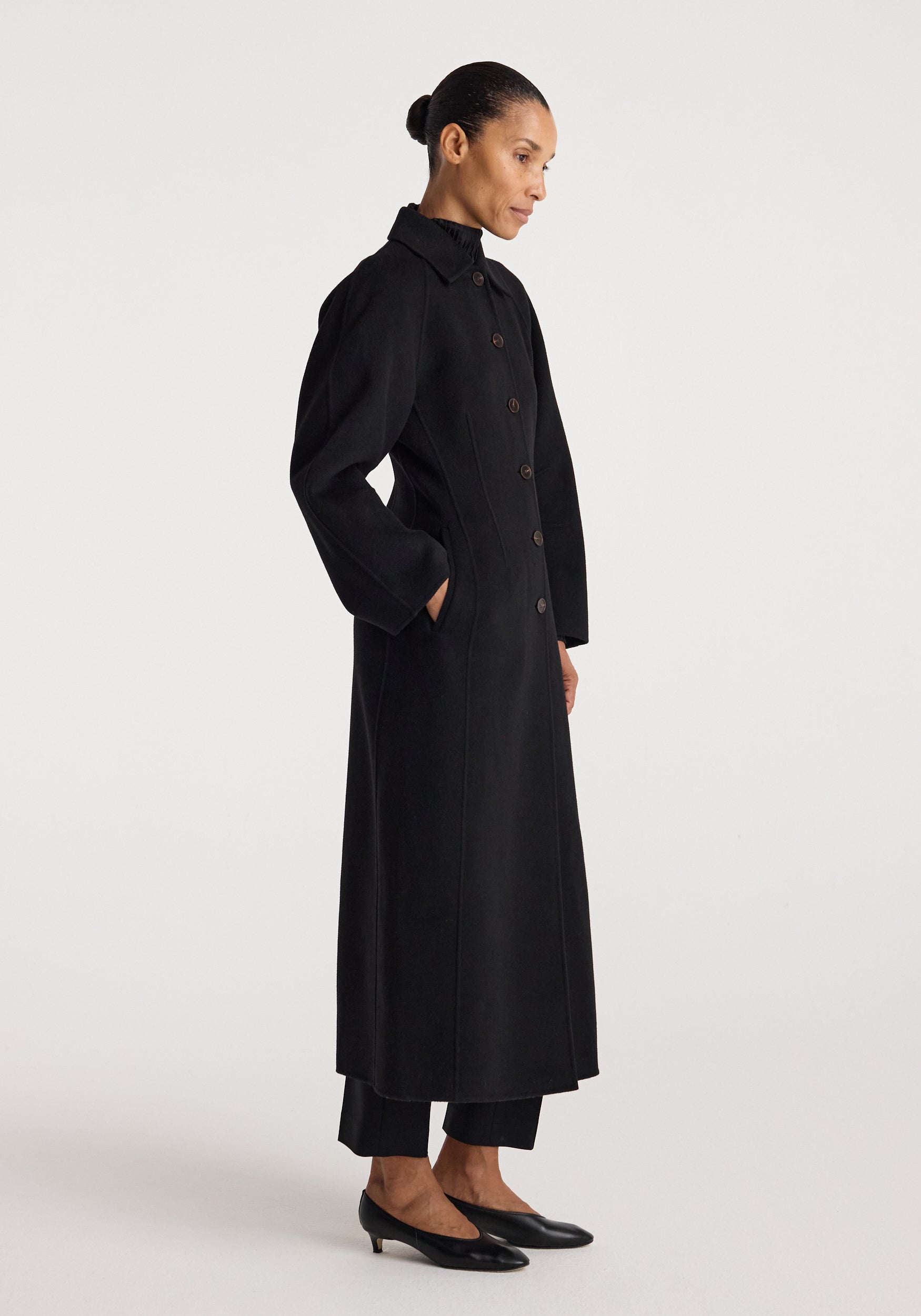 Woman wearing a long black Double-faced elongated sculptural coat | black with button closure, side pockets, and a structured collar, styled with black pants and low heels.