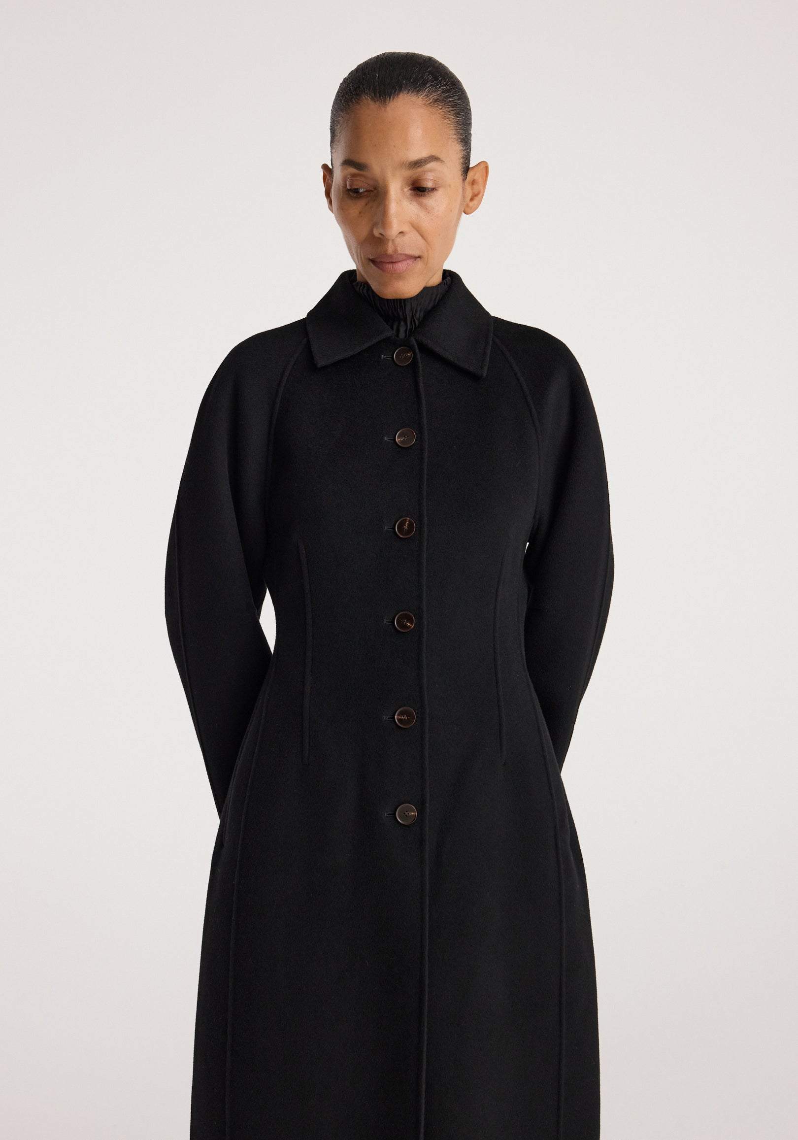 Woman wearing a sleek black Double-faced elongated sculptural coat | black with button closure, structured silhouette, and classic collar, standing against a plain background.