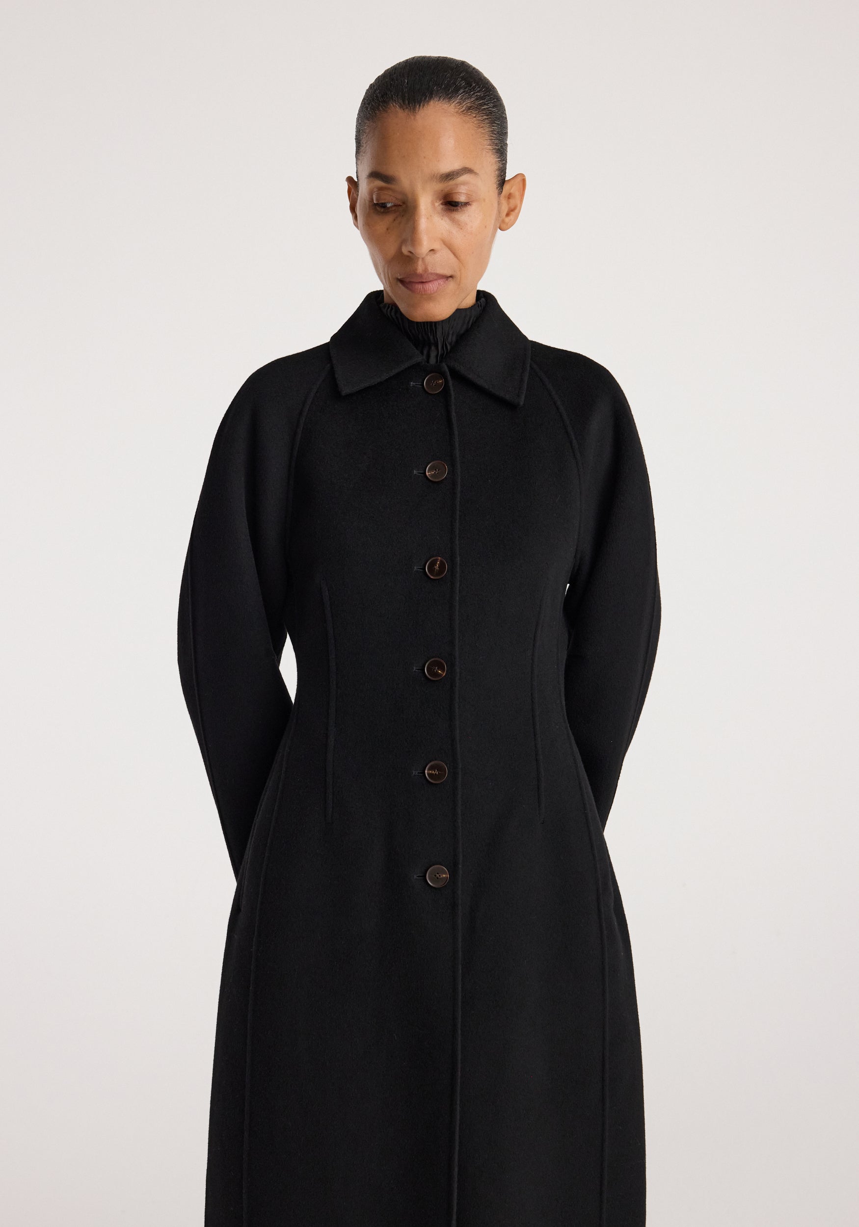 Woman wearing a sleek black Double-faced elongated sculptural coat | black with button closure, structured silhouette, and classic collar, standing against a plain background.