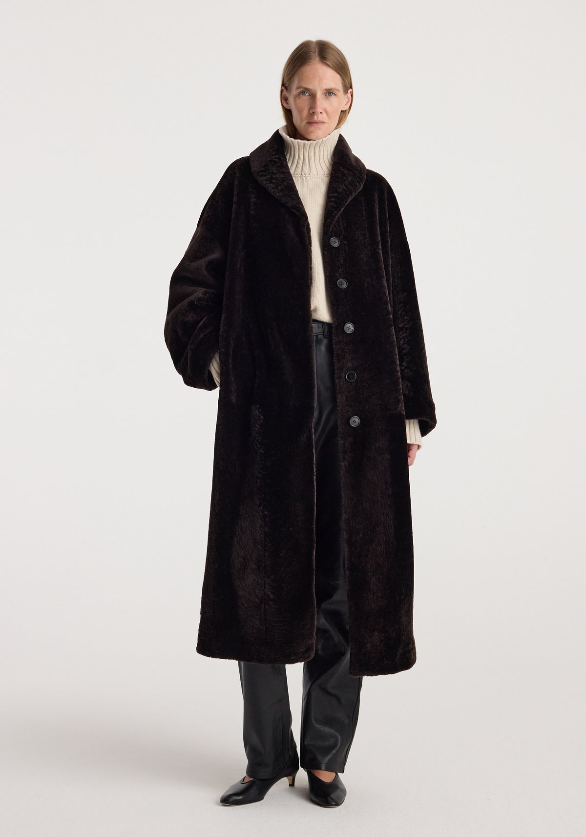 Model wearing the Shearling Shawl-Collar Coat | dark chocolate, a long plush coat styled with a cream turtleneck, dark trousers, and black shoes.