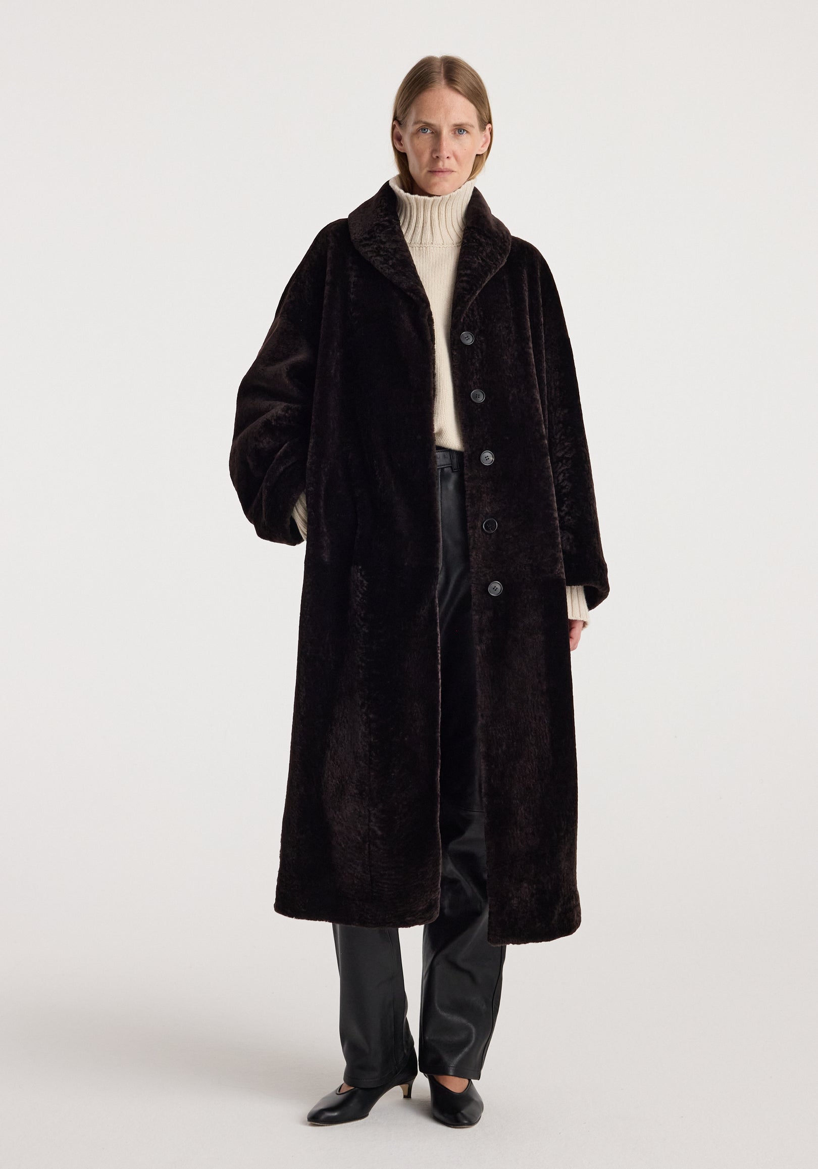 Model wearing the Shearling Shawl-Collar Coat | dark chocolate, a long plush coat styled with a cream turtleneck, dark trousers, and black shoes.