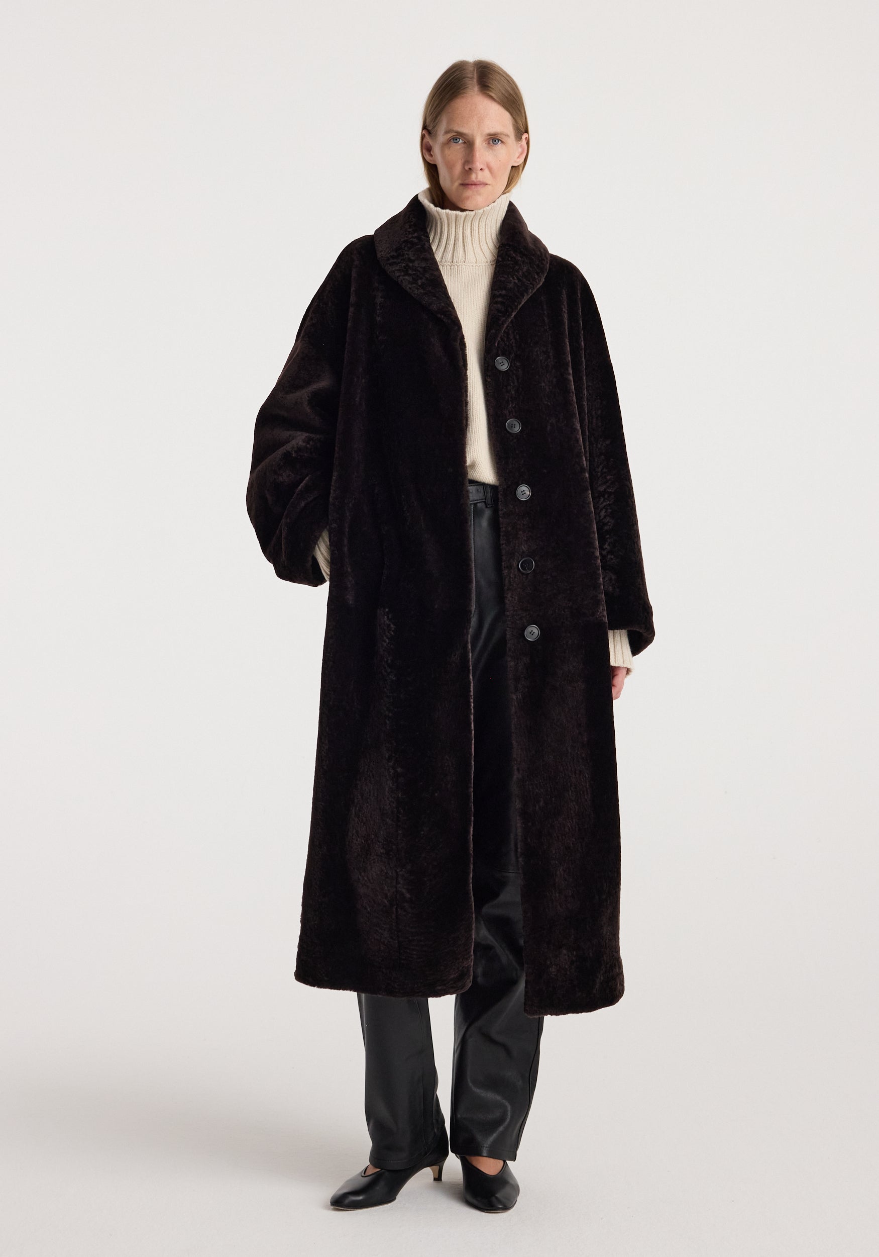 Model wearing the Shearling Shawl-Collar Coat | dark chocolate, a long plush coat styled with a cream turtleneck, dark trousers, and black shoes.