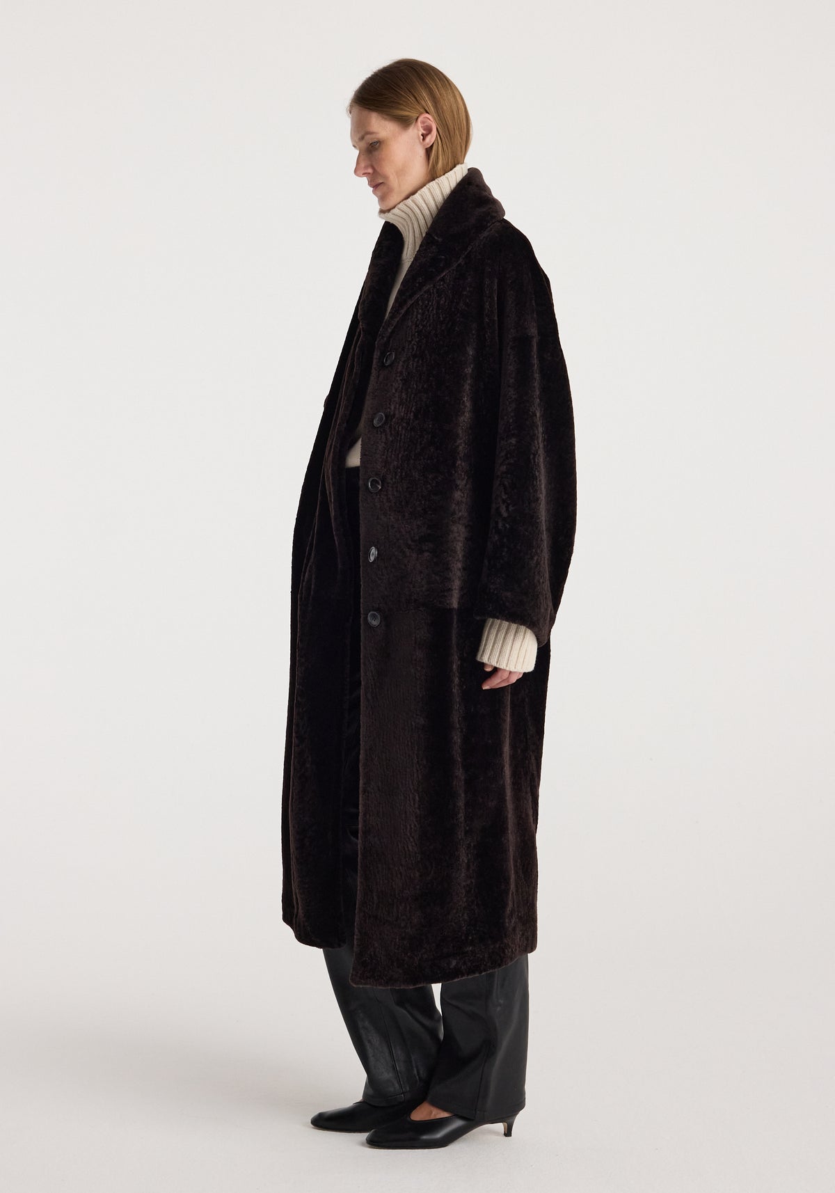Shearling Shawl-Collar Coat | dark chocolate shown on a model in profile with a long dark brown coat, button front, black trousers, and pointed heels