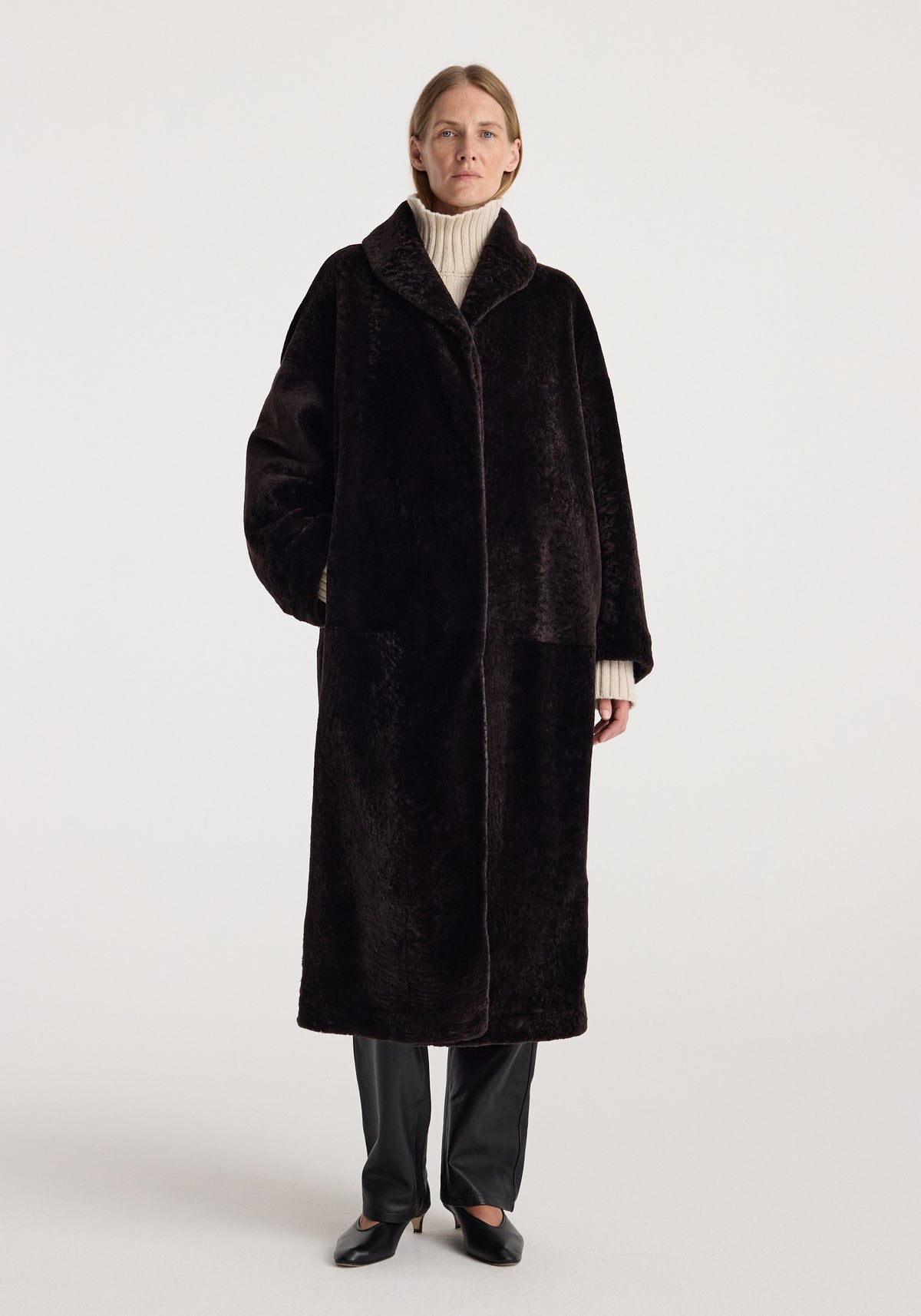 Model wearing a long dark brown shearling coat with a shawl collar over a cream turtleneck, black trousers, and loafers, Shearling Shawl-Collar Coat | dark chocolate