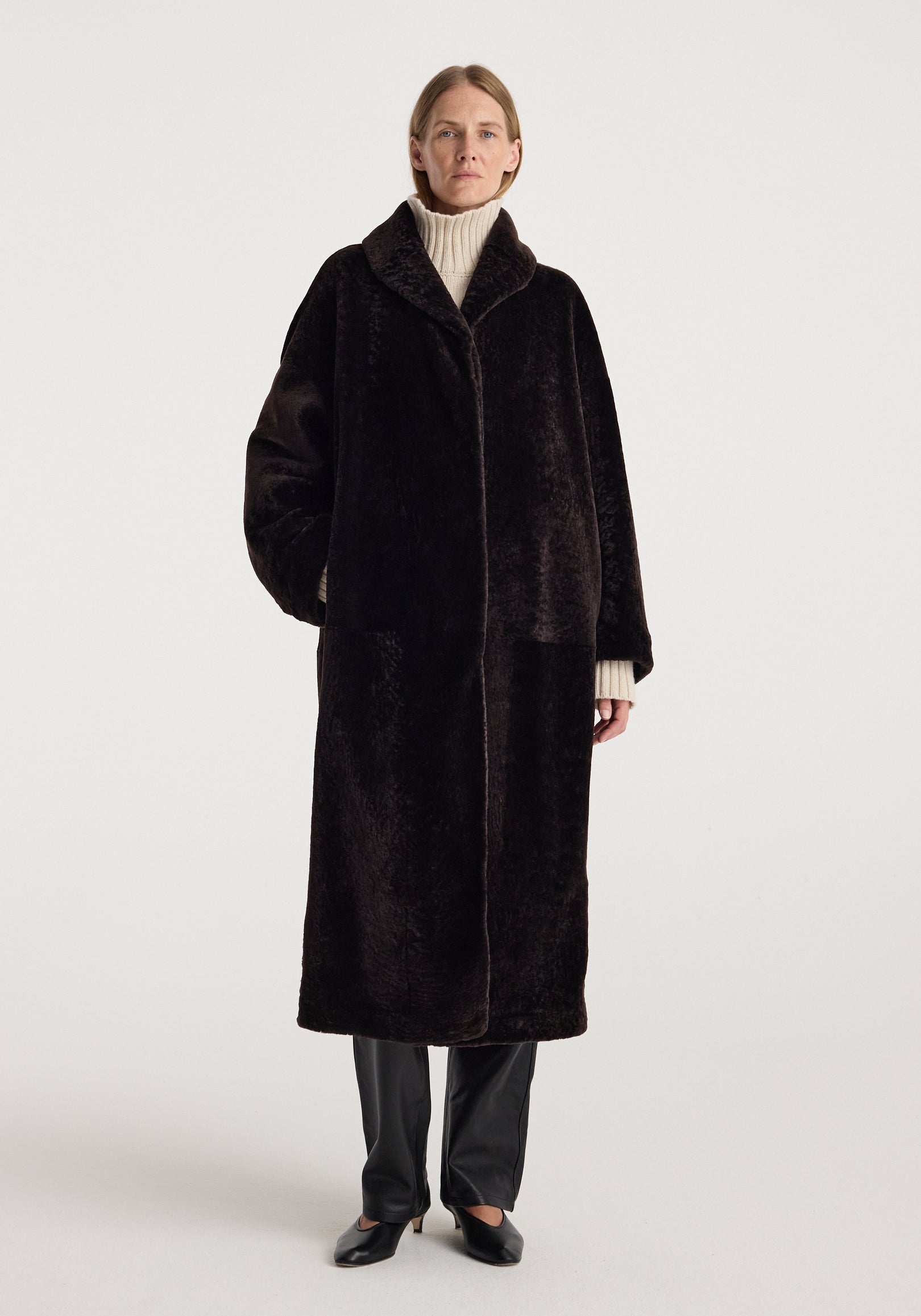 Model wearing a long dark brown shearling coat with a shawl collar over a cream turtleneck, black trousers, and loafers, Shearling Shawl-Collar Coat | dark chocolate