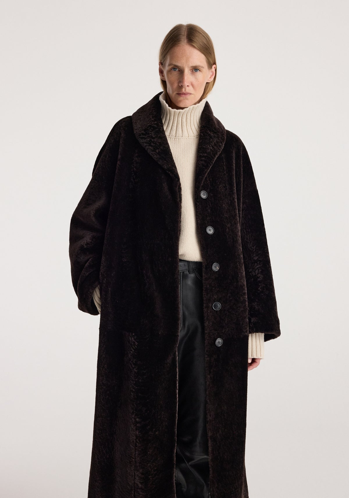 Model wearing a dark chocolate shearling coat with a shawl collar, button front, cream turtleneck sweater, and black trousers. Shearling Shawl-Collar Coat | dark chocolate