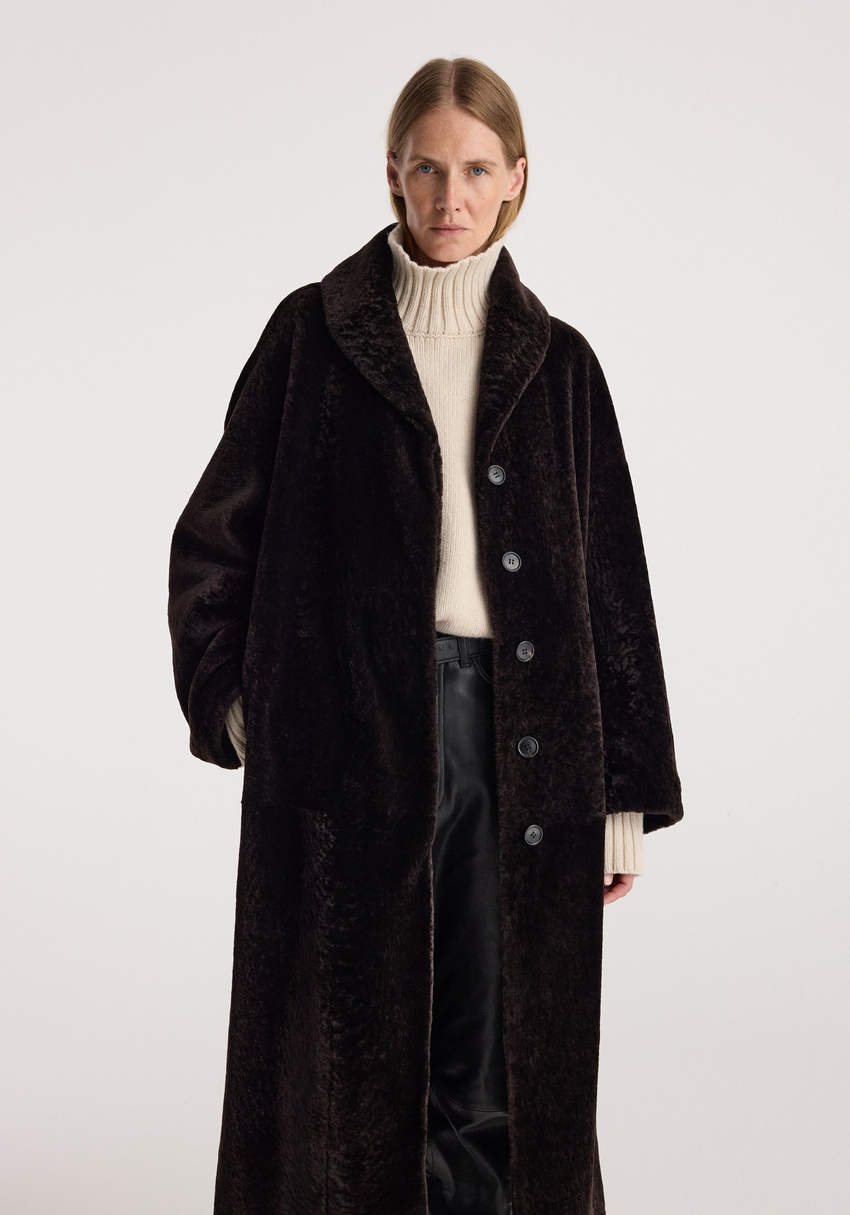 Model wearing a dark chocolate shearling coat with a shawl collar, button front, cream turtleneck sweater, and black trousers. Shearling Shawl-Collar Coat | dark chocolate