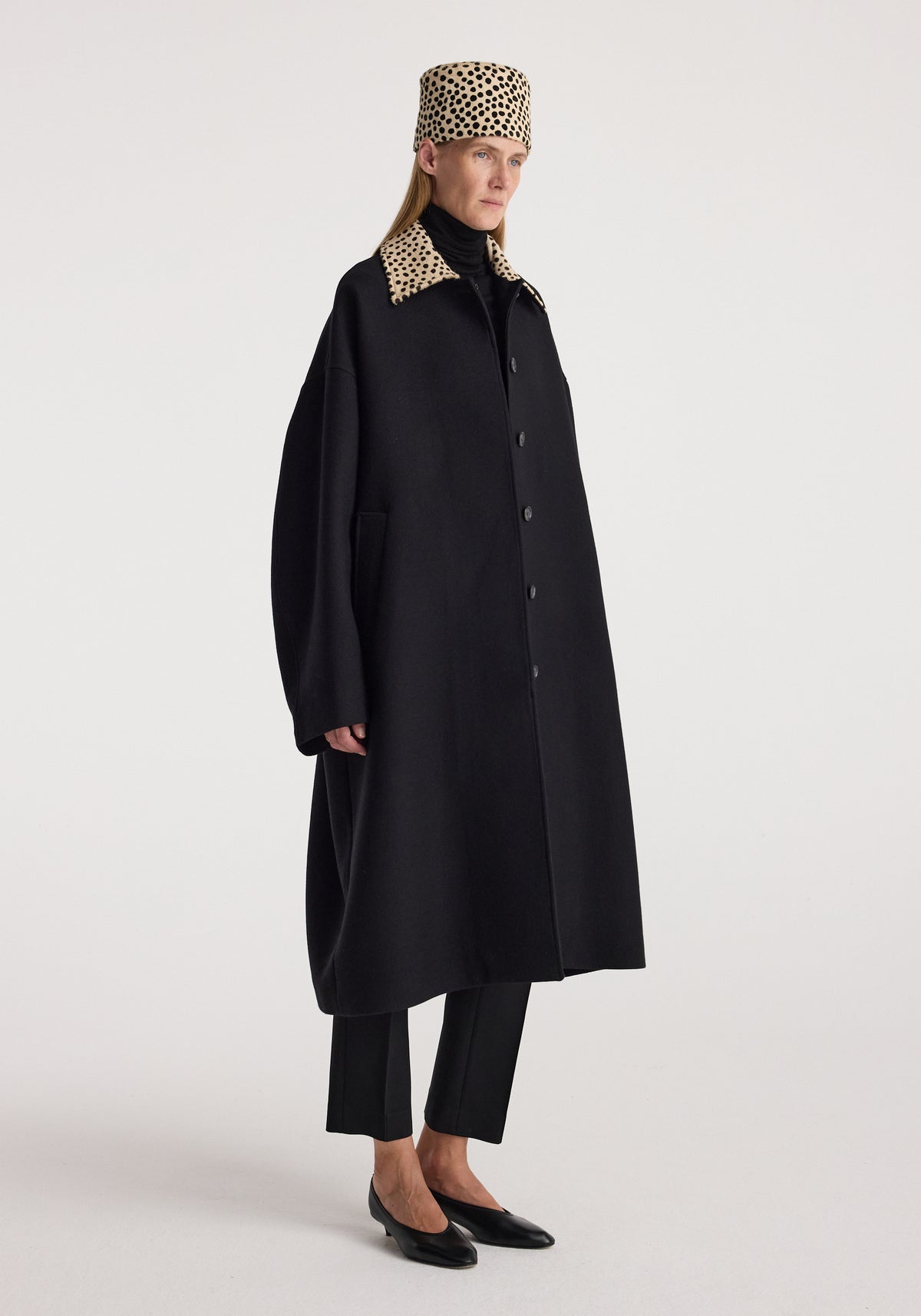 Model wearing the Cheetah collar wool coat | black with a matching cheetah print collar and hat, black trousers, and pointed black flats against a neutral background