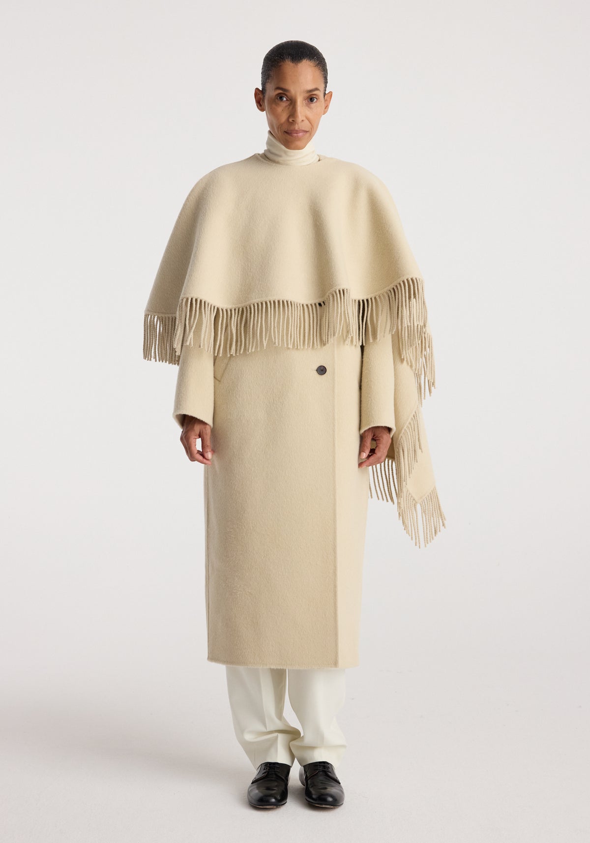 Model wearing a long cream coat with fringe details over shoulders, paired with white pants and black shoes, showcasing the Double-faced fringe coat | butter.