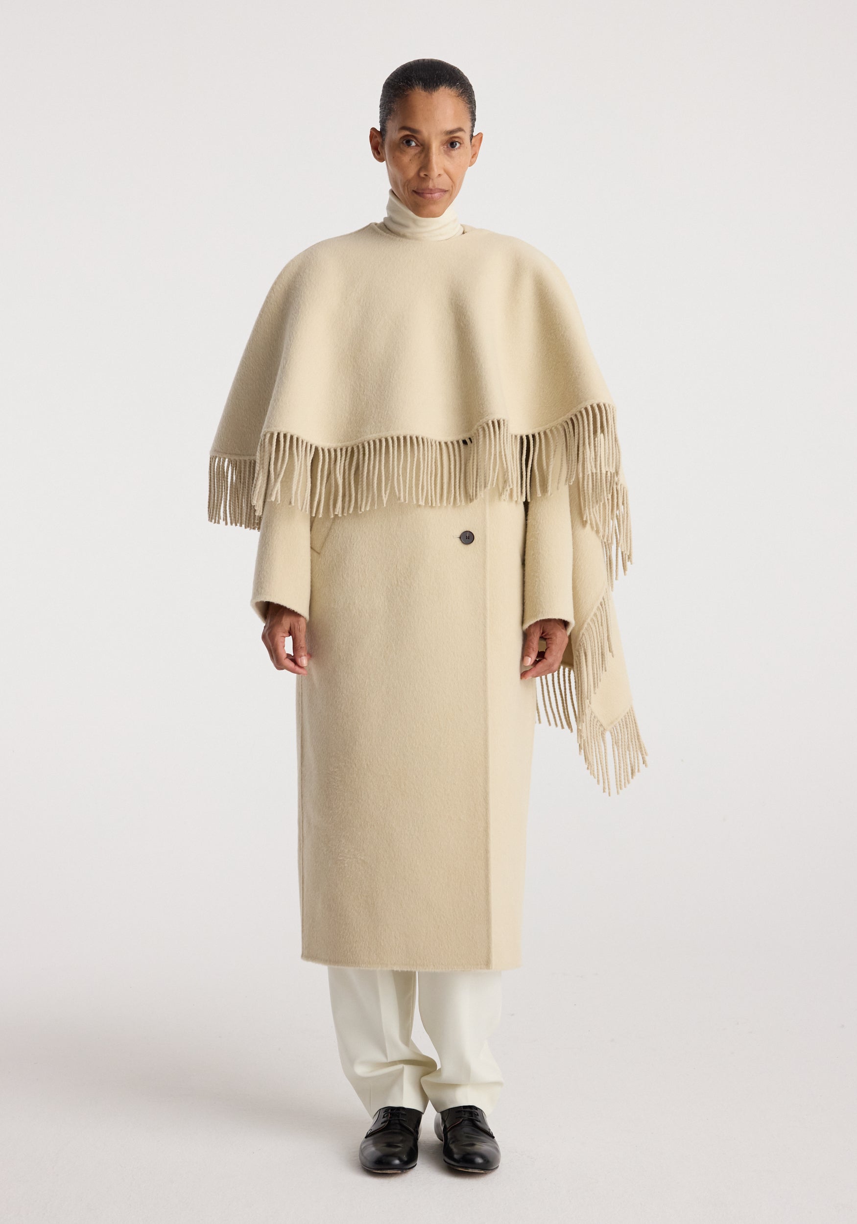 Model wearing a long cream coat with fringe details over shoulders, paired with white pants and black shoes, showcasing the Double-faced fringe coat | butter.