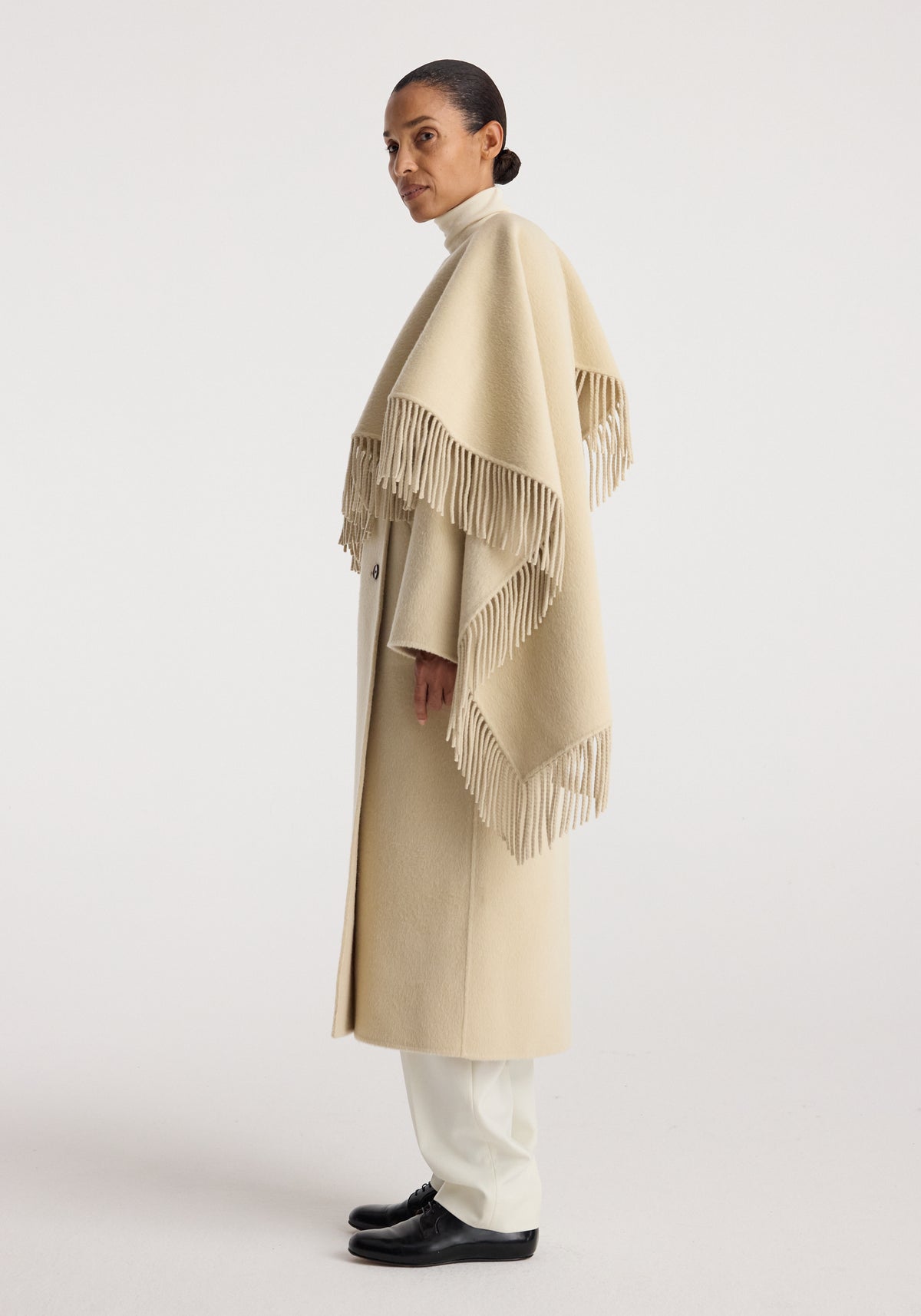 Profile view of a woman wearing a long beige double-faced fringe coat | butter with a turtleneck and white pants, paired with black shoes, against a plain background.
