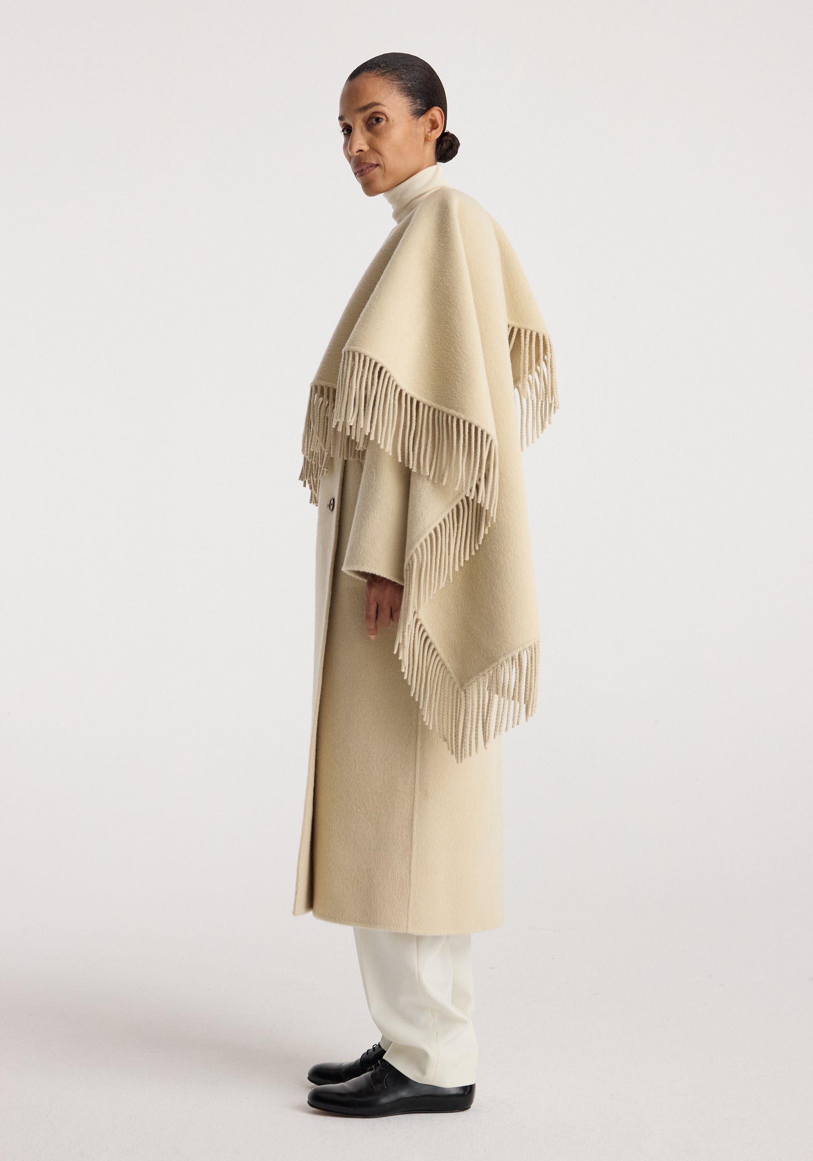 Profile view of a woman wearing a long beige double-faced fringe coat | butter with a turtleneck and white pants, paired with black shoes, against a plain background.