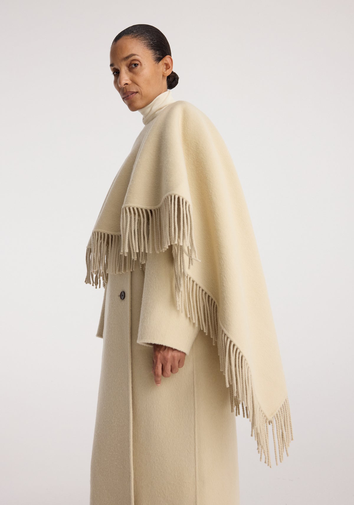 Woman wearing a warm, elegant Double-faced fringe coat | butter in a soft cream color with long fringe details along the edges, showcasing a sophisticated minimalist style.