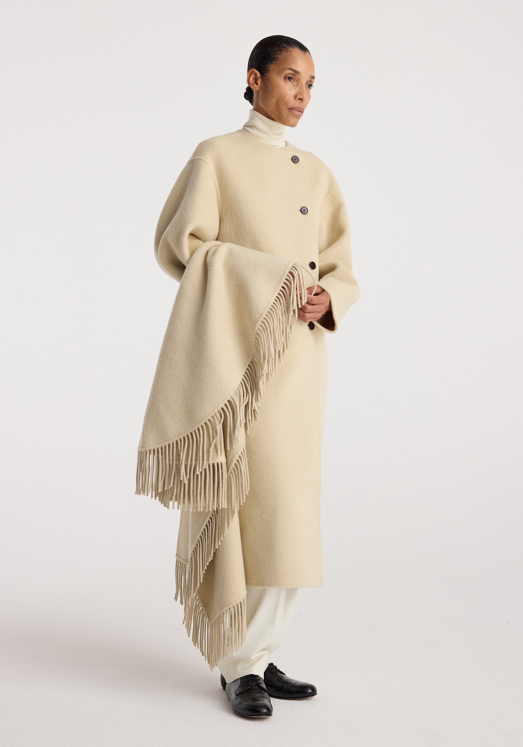 Woman wearing a cream-colored Double-faced fringe coat | butter with large buttons and long fringed edges, styled with white pants and black shoes against a neutral background.
