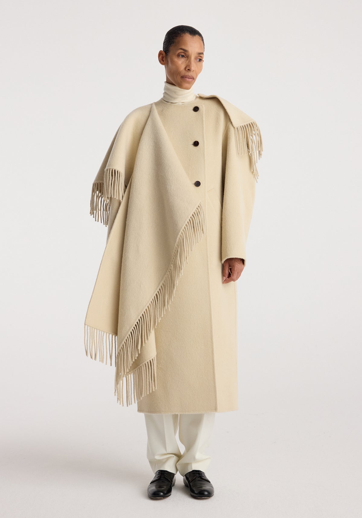 Woman wearing a cream-colored long coat with fringe details and buttons, styled over white pants and a turtleneck, showcasing the Double-faced fringe coat | butter.