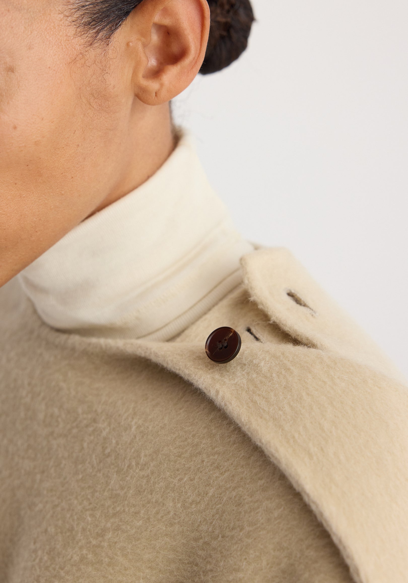 Close-up of a person wearing a soft, textured butter-colored Double-faced fringe coat | butter with a dark button detail on the shoulder over a cream turtleneck.