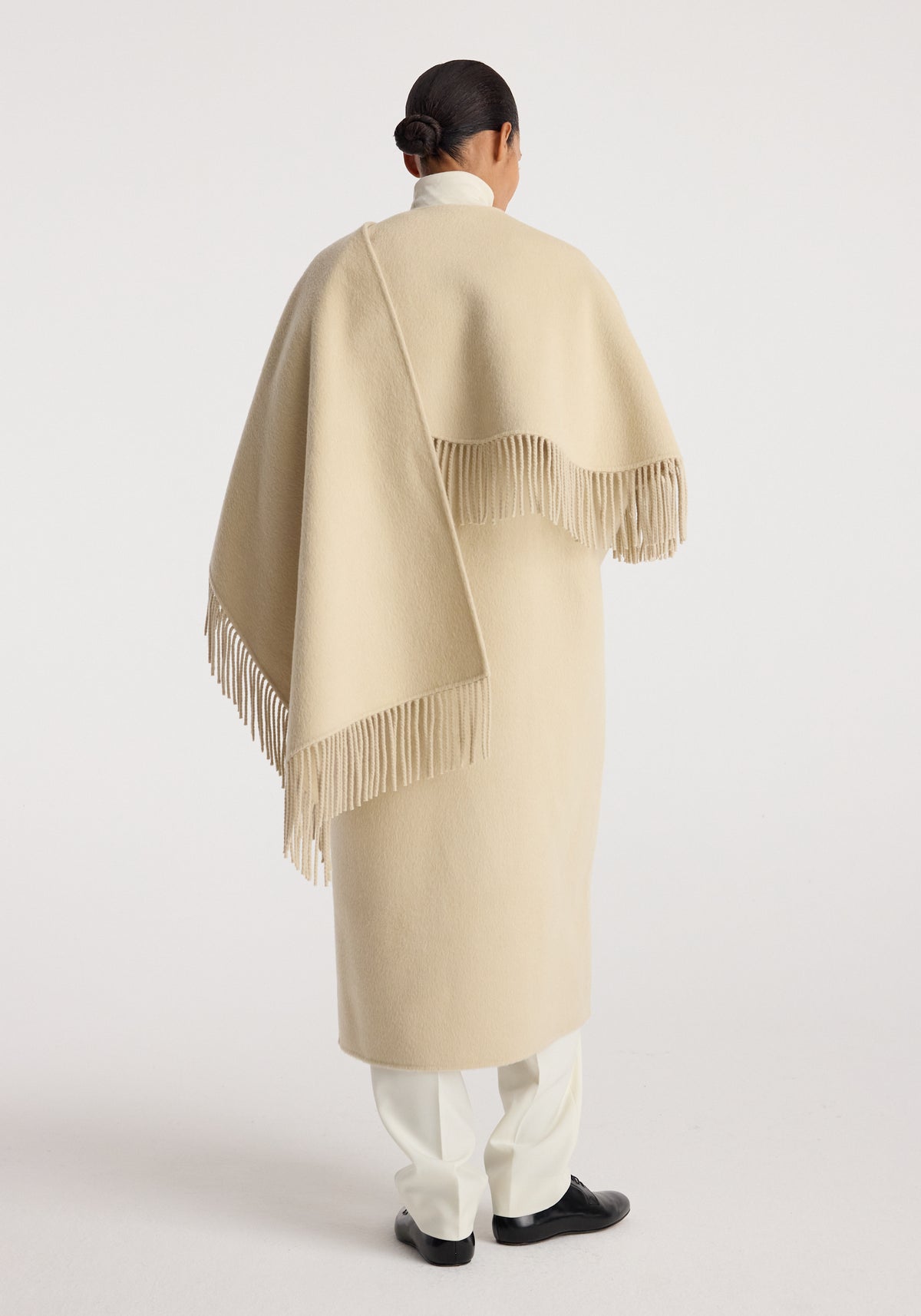 Woman seen from behind wearing a cream-colored double-faced fringe coat | butter with fringe detailing along the edges, paired with white pants and black shoes.