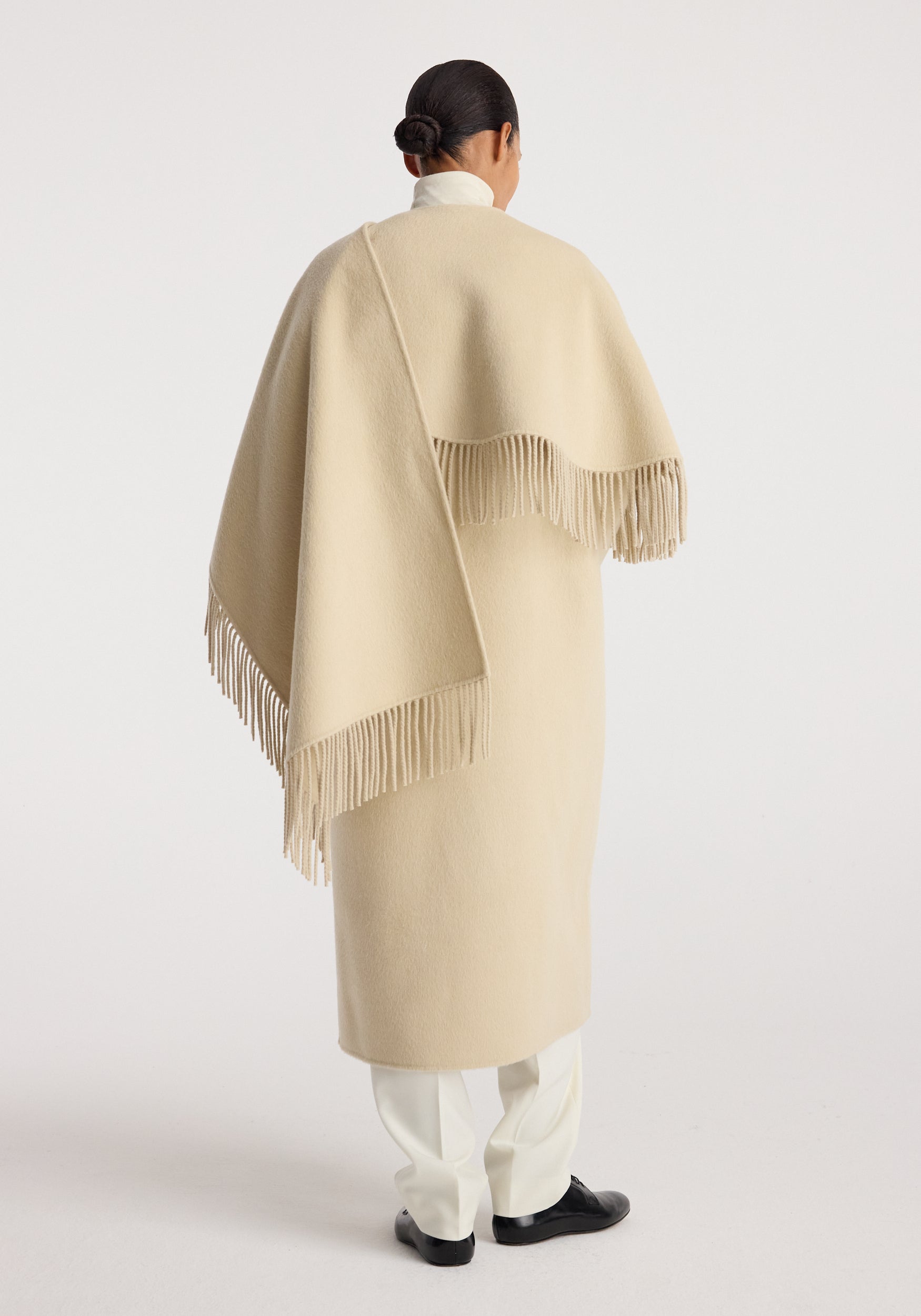 Woman seen from behind wearing a cream-colored double-faced fringe coat | butter with fringe detailing along the edges, paired with white pants and black shoes.