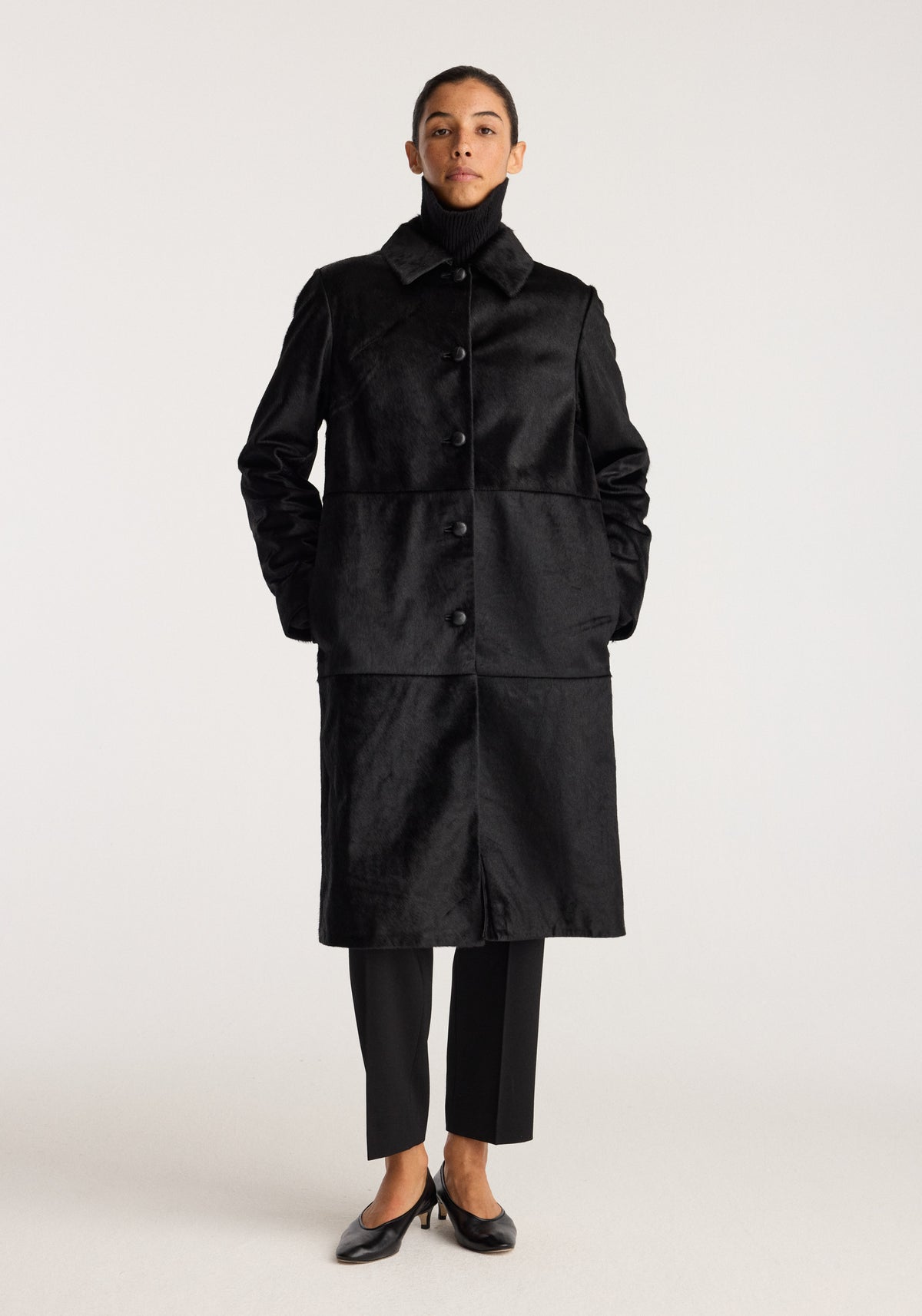 Black ponyhair coat | black shown on a model with a button front collar side pockets and knee length silhouette styled with black trousers and loafers