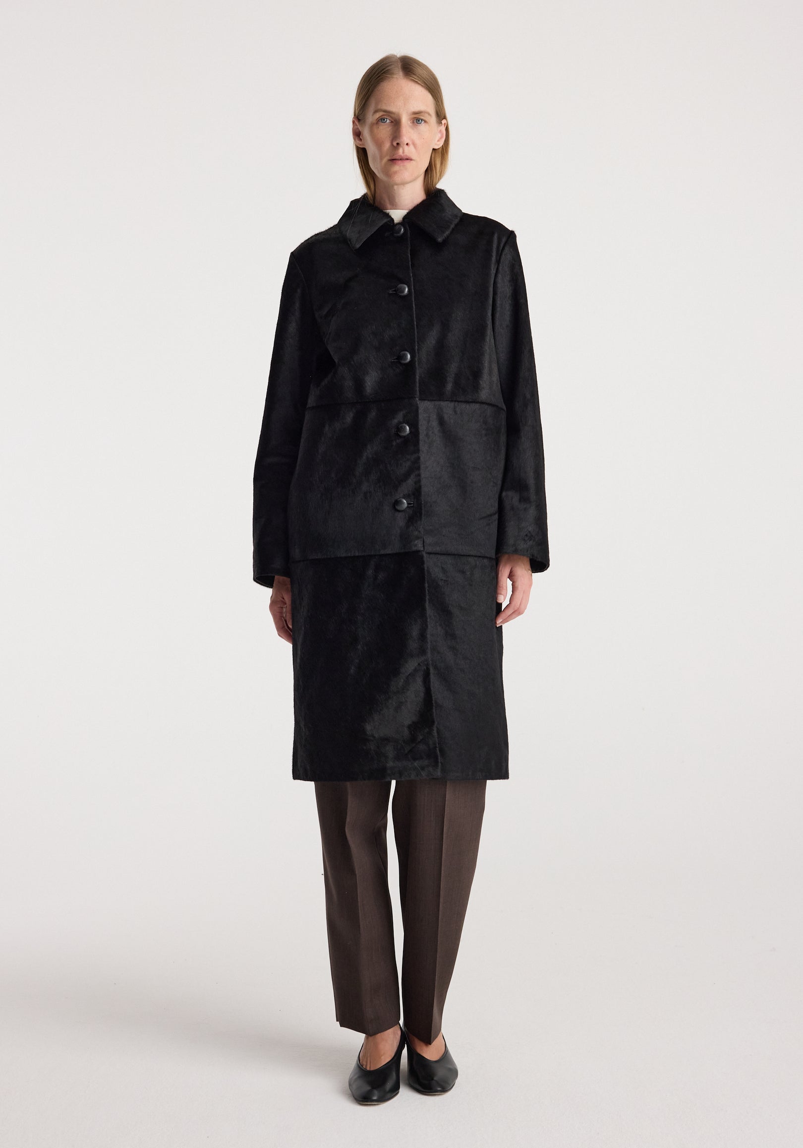 Woman wearing Black ponyhair coat | black, buttoned front and knee-length design with long sleeves and collar, paired with dark trousers and black shoes.
