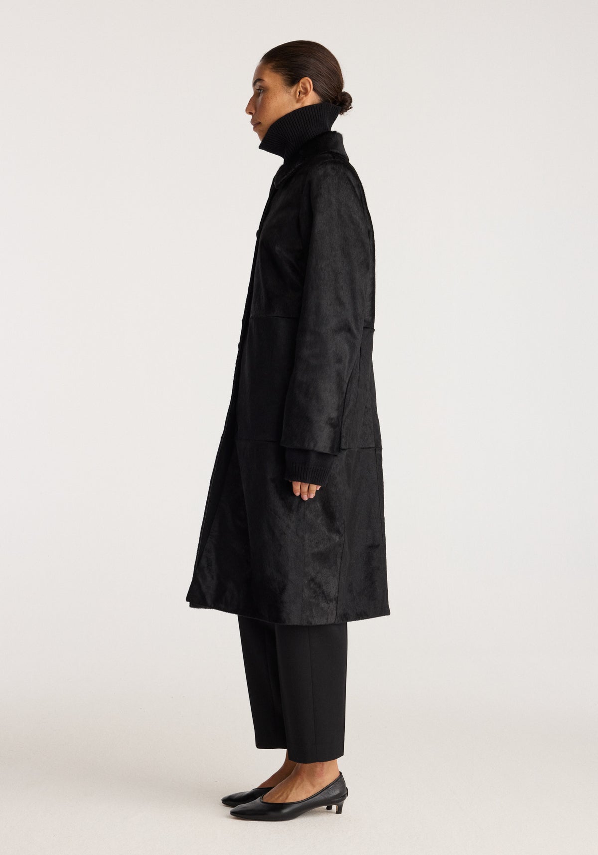 Black ponyhair coat | black shown in side view on a model with a high collar long sleeves and a knee length silhouette styled with black trousers and heels