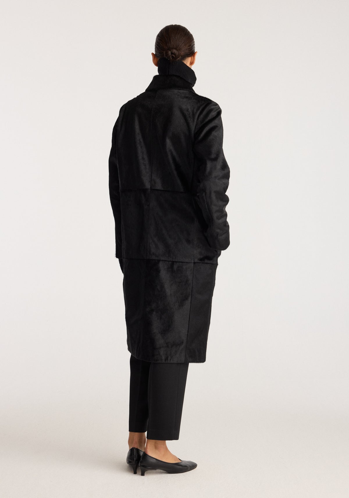 Black ponyhair coat | black shown from the back on a model with a high collar long sleeves and knee length fit