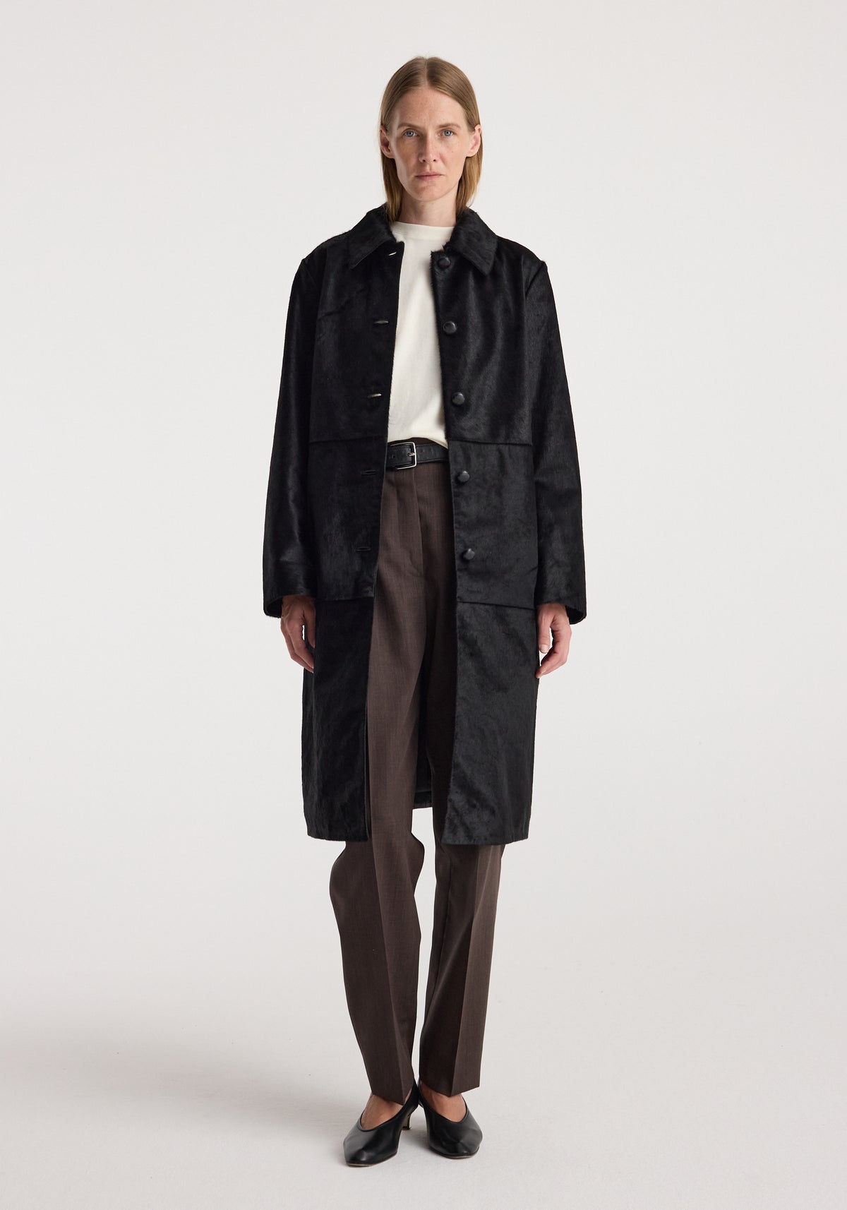Woman wearing a long black ponyhair coat | black with buttons, paired with brown pants, black shoes, and a white top, standing against a white background.
