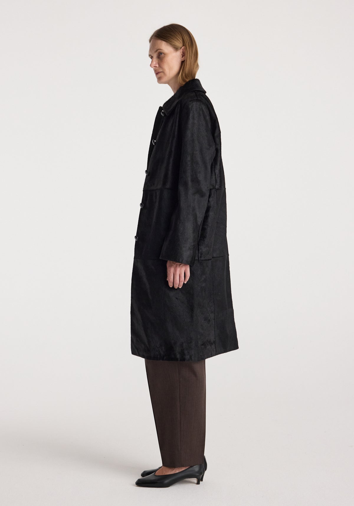 Side profile of a woman wearing a Black ponyhair coat | black with long sleeves, paired with brown pants and black heels against a plain background.