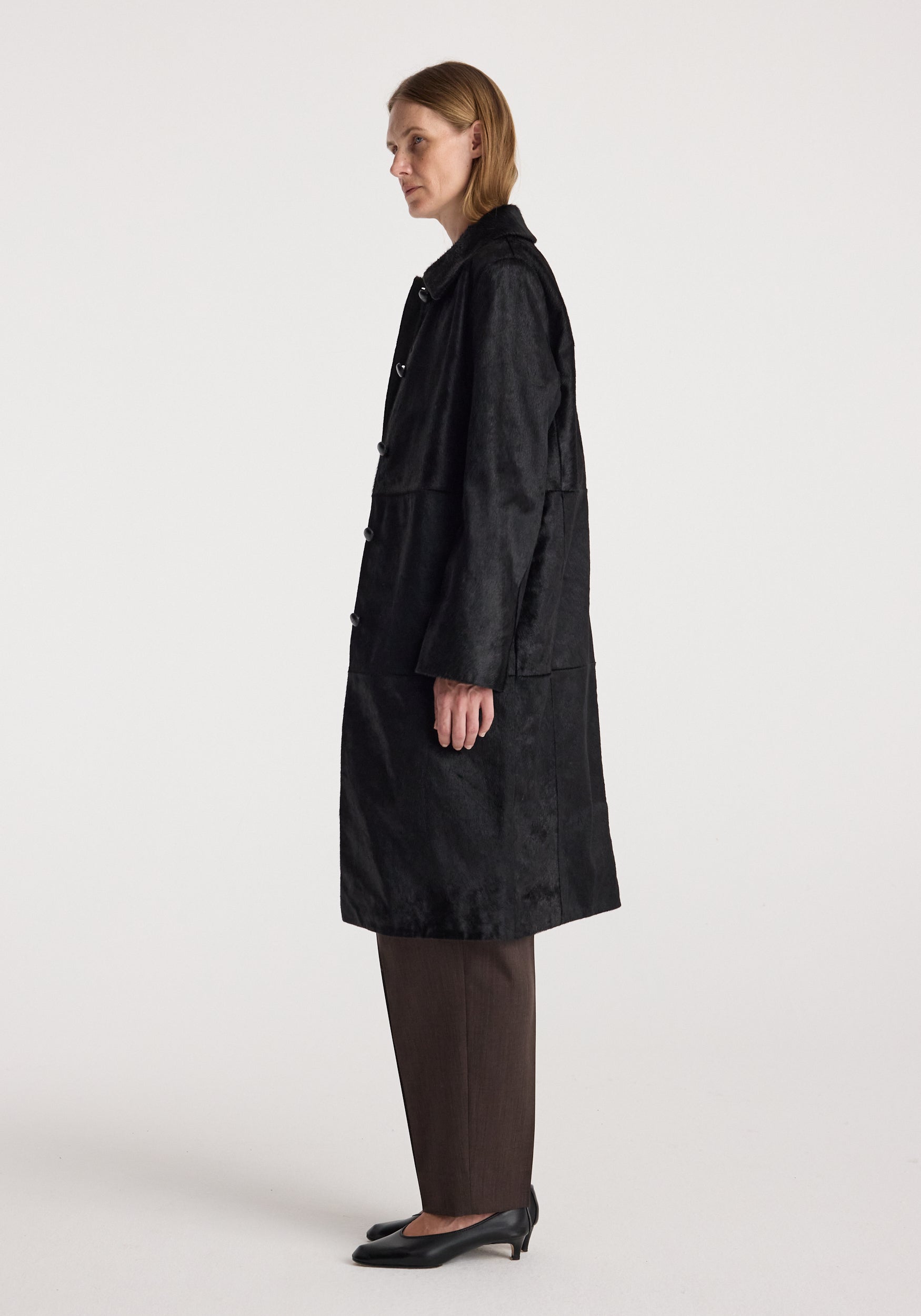 Side profile of a woman wearing a Black ponyhair coat | black with long sleeves, paired with brown pants and black heels against a plain background.