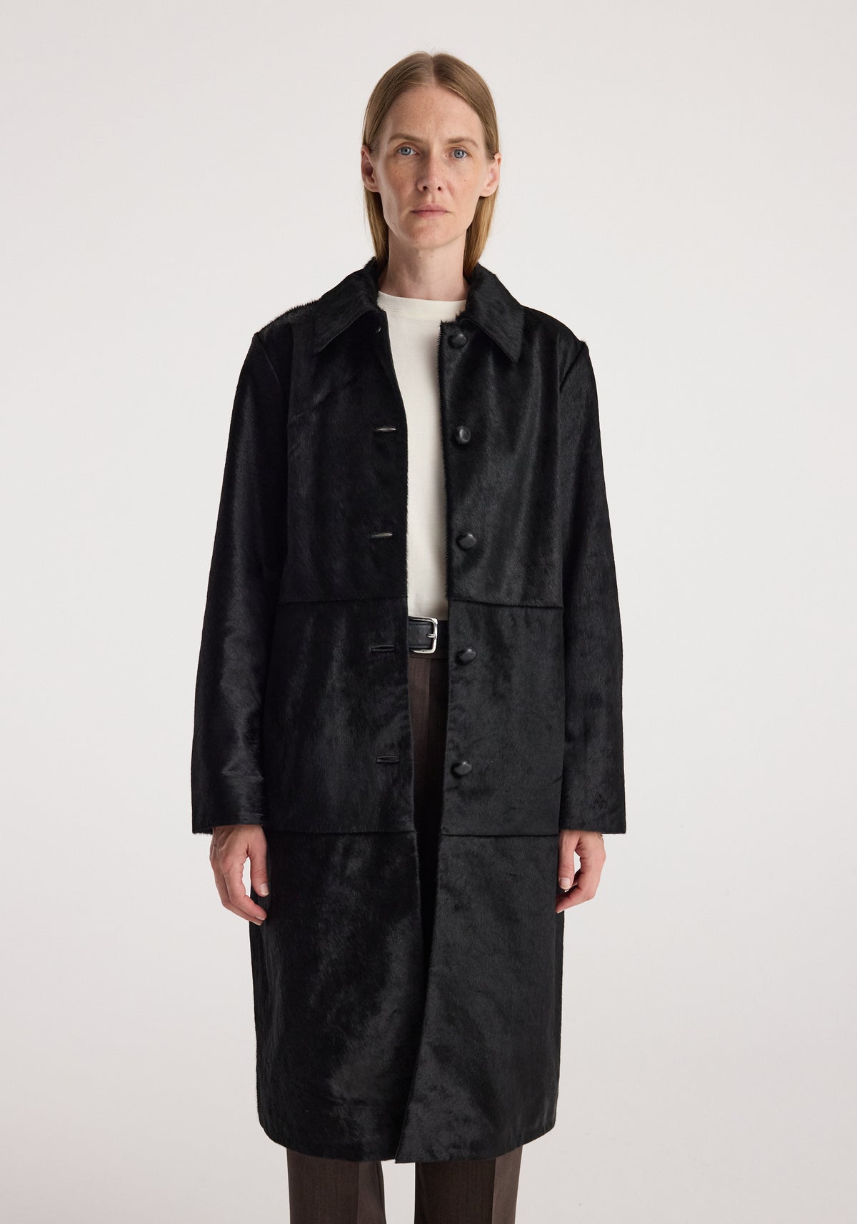 Woman wearing a black ponyhair coat | black with a button-up front and a classic collar, styled over a white top and dark pants against a plain background.